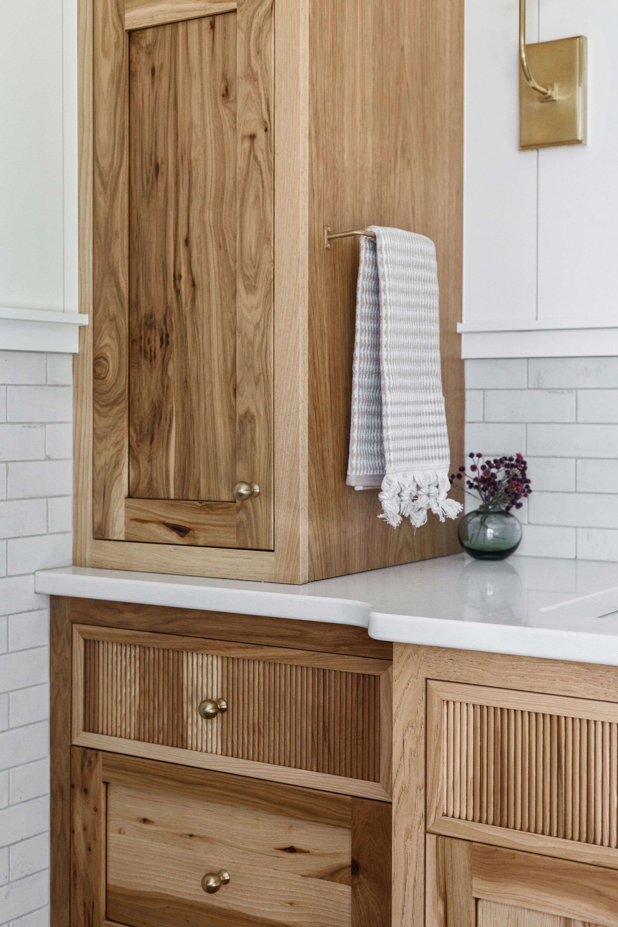 Detailed view of a custom wood cabinet with a towel hanging on a gold bar, positioned above a countertop and featuring reeded drawers. The background includes white subway tiles.