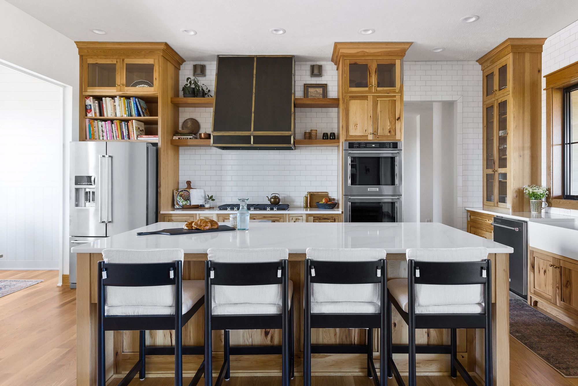 Spacious kitchen features natural wood cabinetry, a large white countertop island with seating, and stainless steel appliances. A black range hood and subway tile backsplash add modern touches.
