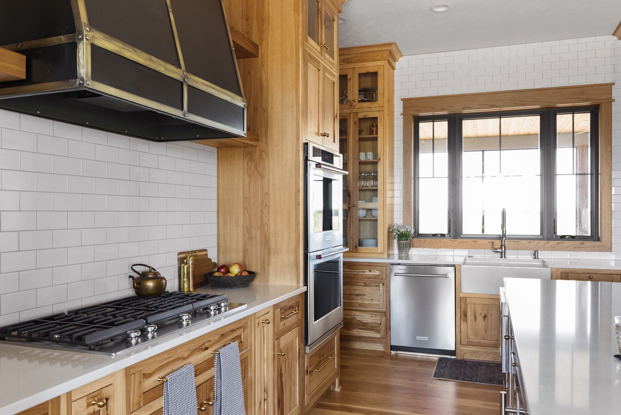 Bright kitchen with white subway tile backsplash and warm wood cabinetry. Stainless steel appliances including a built-in oven and a large window above a farmhouse sink.