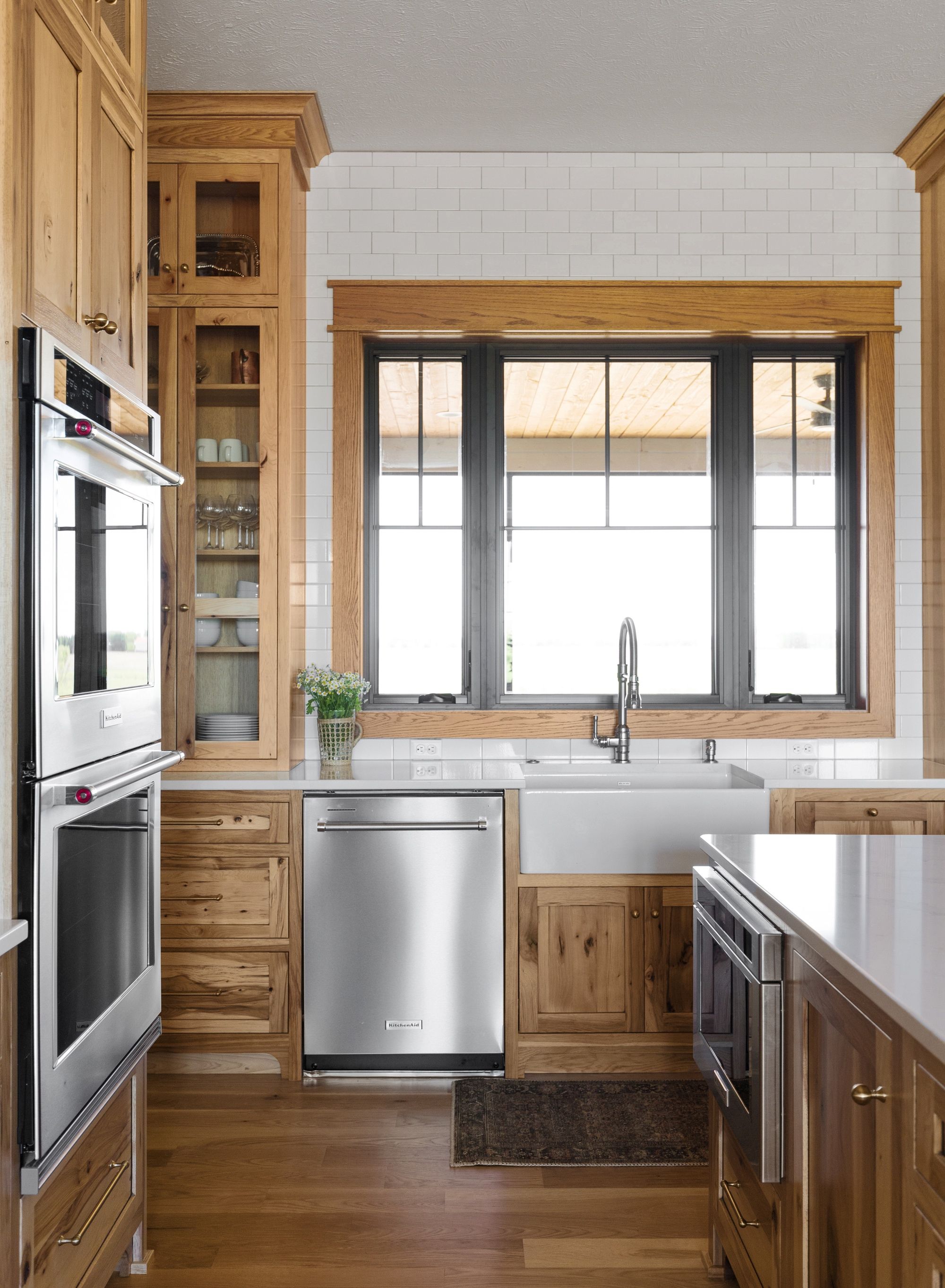 Bright kitchen with natural wood cabinetry, stainless steel appliances, and a white farmhouse sink. A large black-framed window provides ample natural light, complemented by white subway tile backsplash.