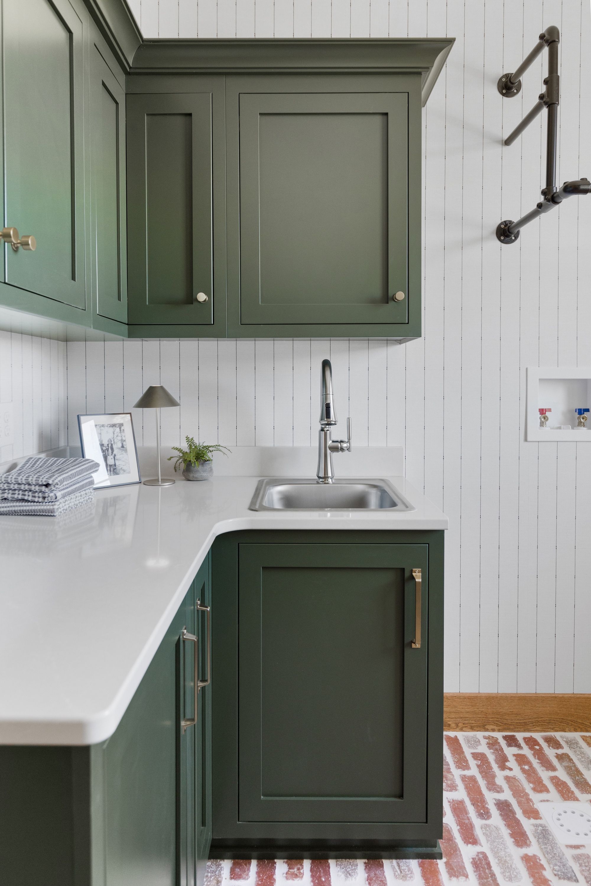 Laundry room featuring deep green cabinets, white countertops, and a stainless steel sink with a modern faucet. A decorative plumbing fixture and patterned wallpaper add visual interest to the space.