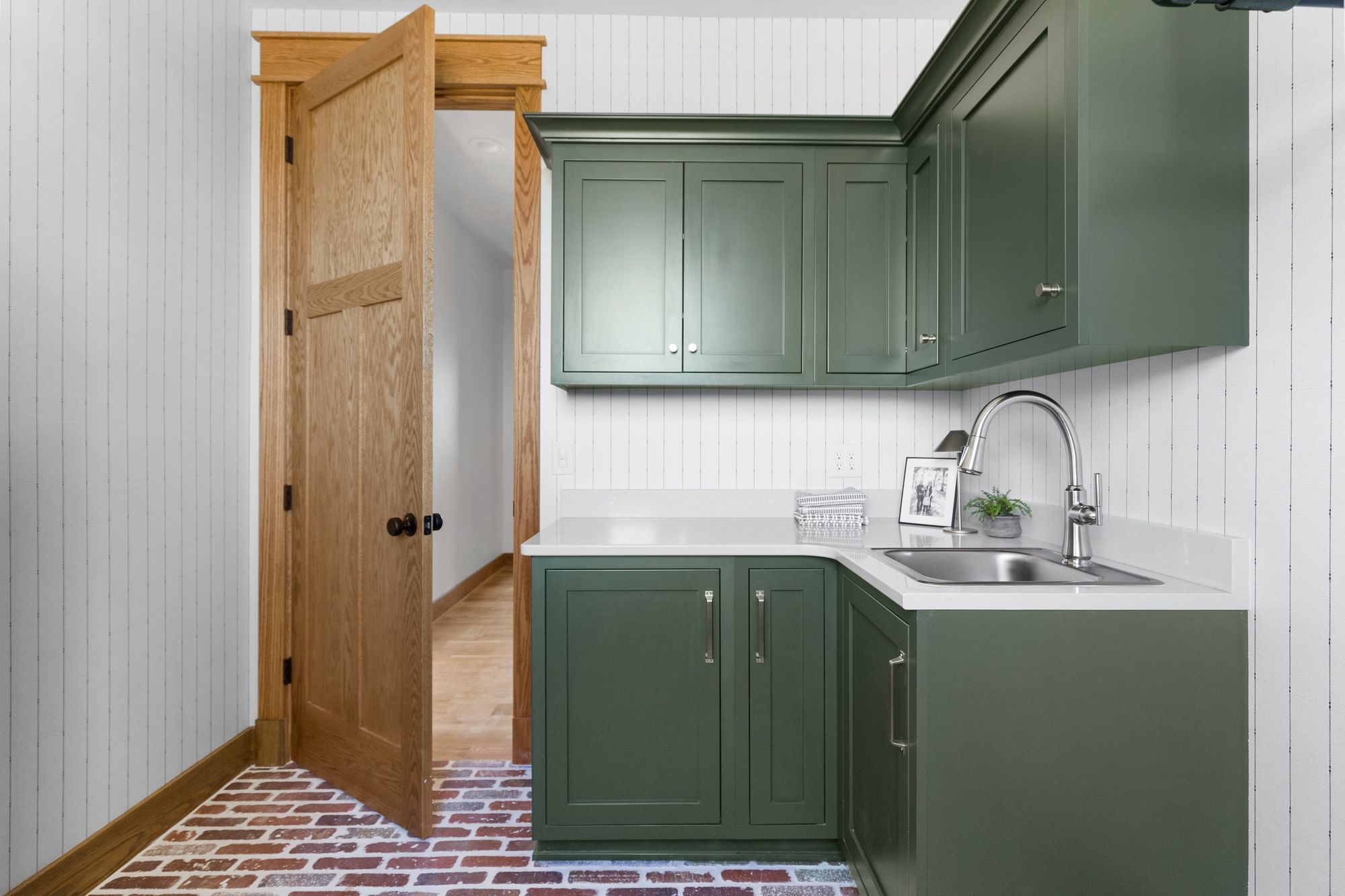 Laundry room featuring green cabinets, white countertops, and brick floors. An open wooden door leads to a hallway.