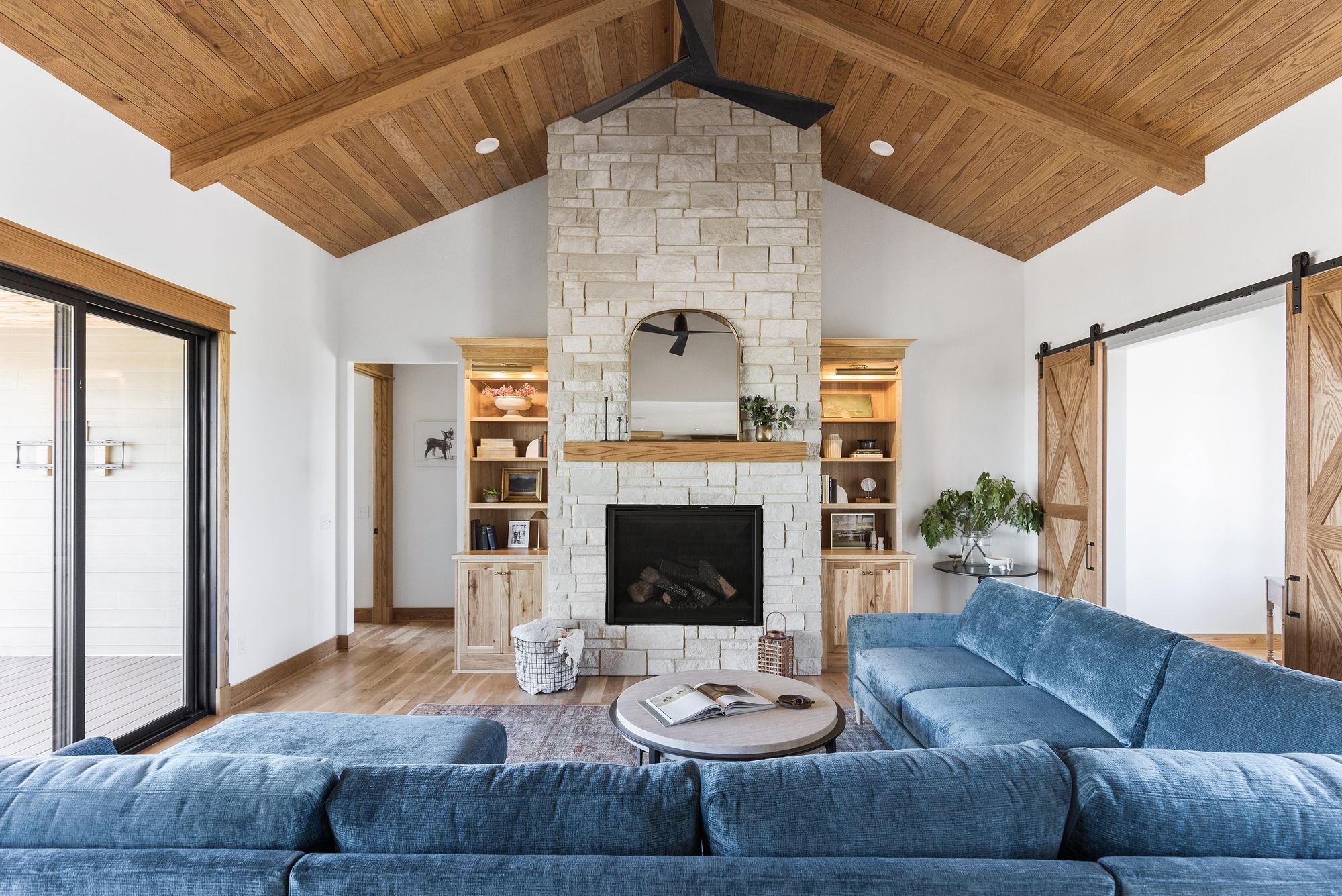 Bright living room features a stone fireplace flanked by built-in shelves, a blue sectional sofa, and a round coffee table. Wood ceiling and sliding barn doors add rustic warmth to the space.