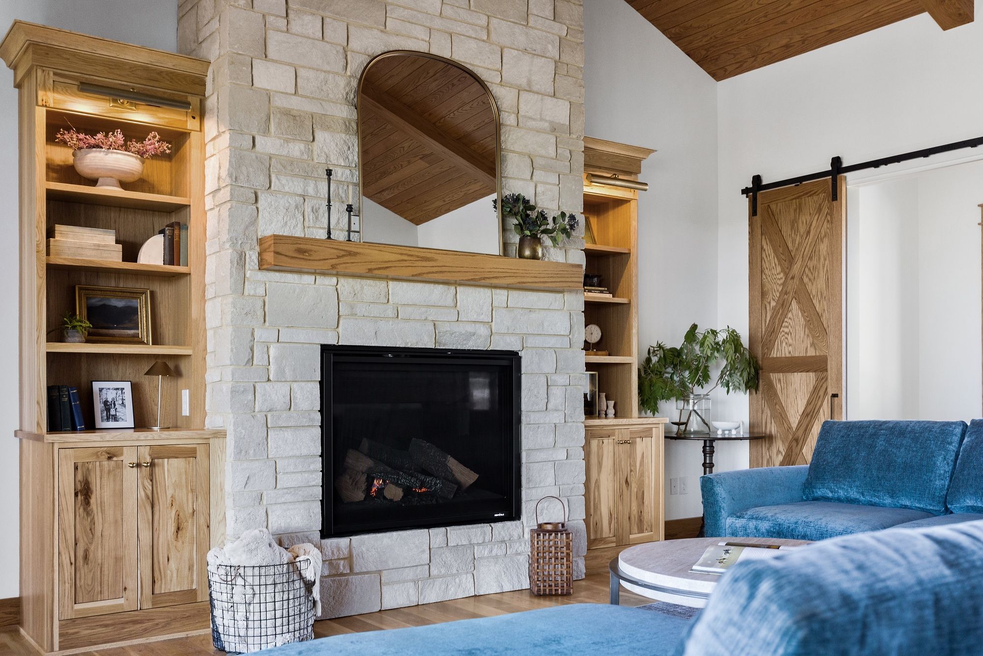 Living room featuring a stone fireplace with built-in wooden shelving on either side and a wooden mantel. The fireplace is topped with an arched mirror reflecting a wooden ceiling. A blue sofa and sliding barn door are also visible.