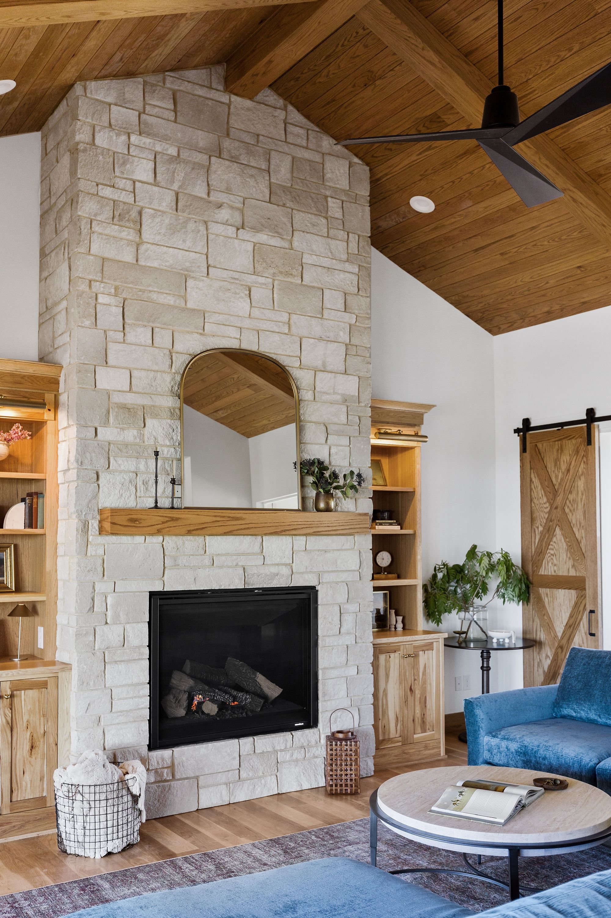 Bright living room with stone fireplace and built-in shelving. The room features a vaulted wood ceiling, a barn door, and a blue sectional sofa, creating a cozy and modern atmosphere.