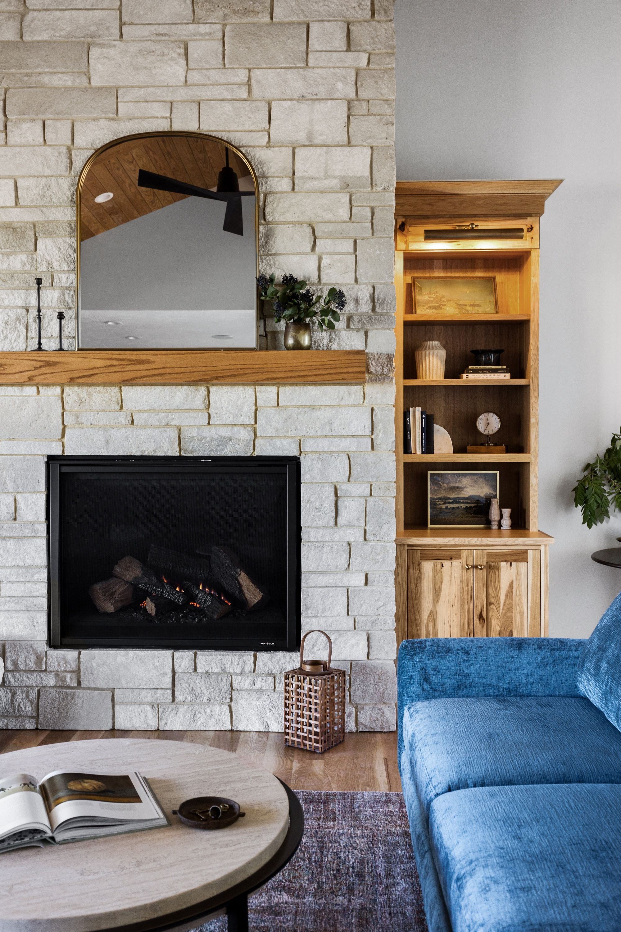 Living room features a stone fireplace with a black mesh screen and wood mantel, beside a built-in oak bookshelf. The room includes a round coffee table and a blue velvet sofa, complemented by a patterned rug.