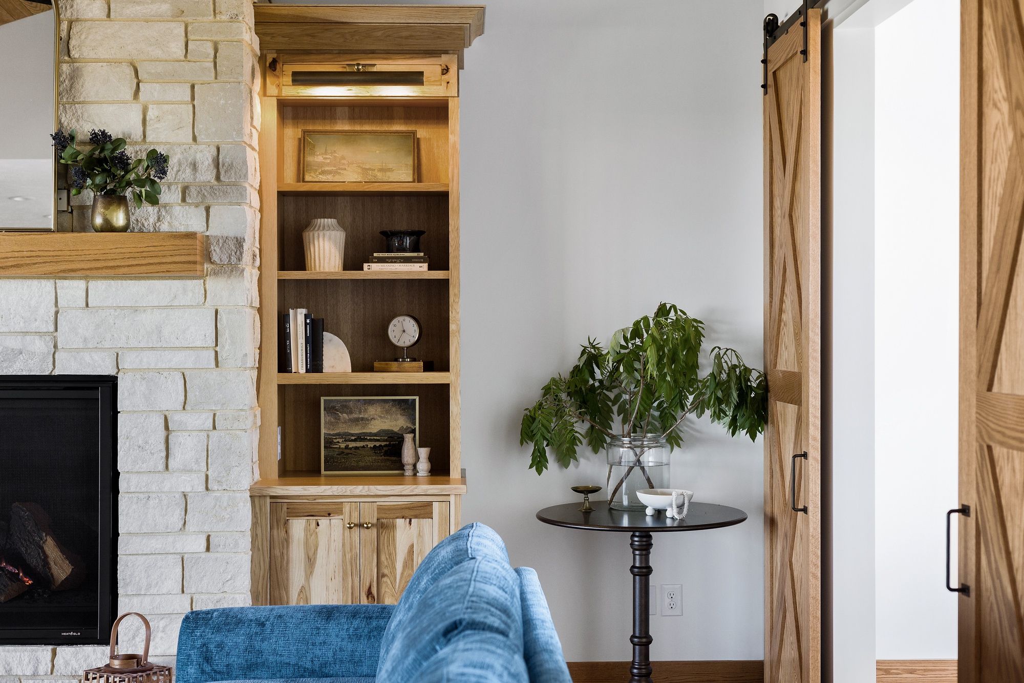 Cozy living room featuring a stone fireplace with built-in shelving filled with decorative items. A round side table displays plants next to a sliding barn door.