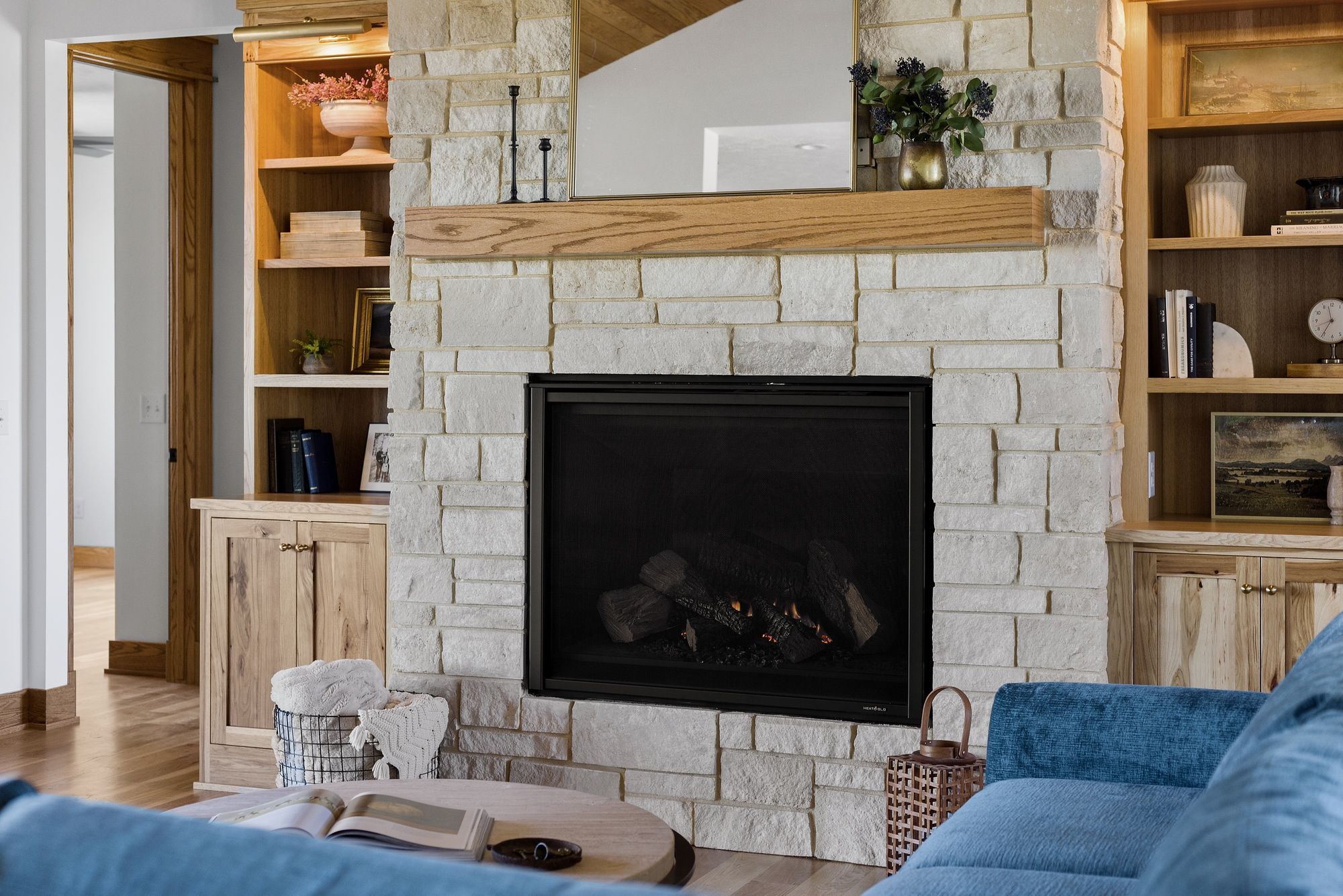 The image depicts a living room with a stone fireplace as its focal point. The fireplace is flanked by built-in wooden shelves and cabinets, complemented by a wooden mantelpiece and a decorative mirror above the fireplace.