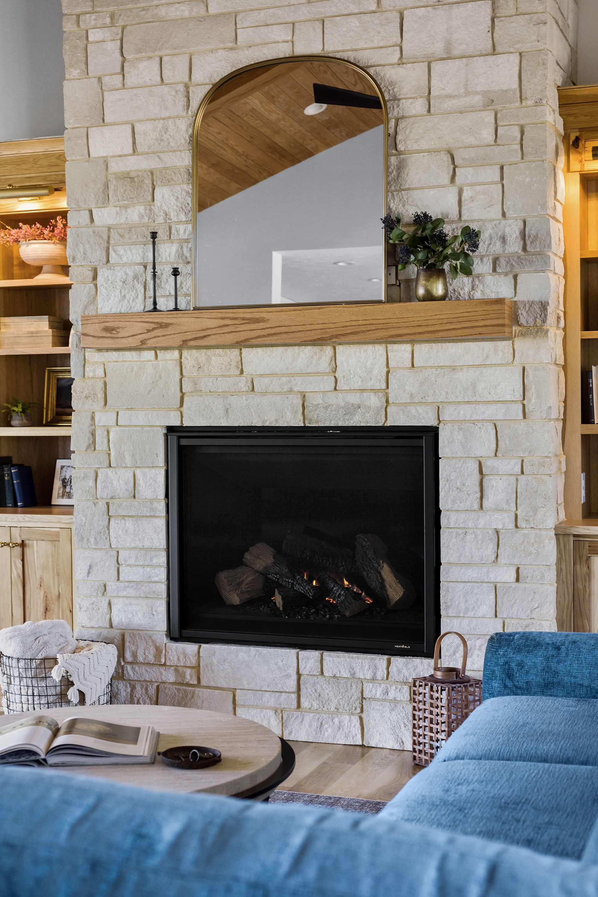 The living room features a stone fireplace with a wooden mantel, a gold-framed arched mirror, and built-in shelving on either side. The room is furnished with a round coffee table and a blue sofa.