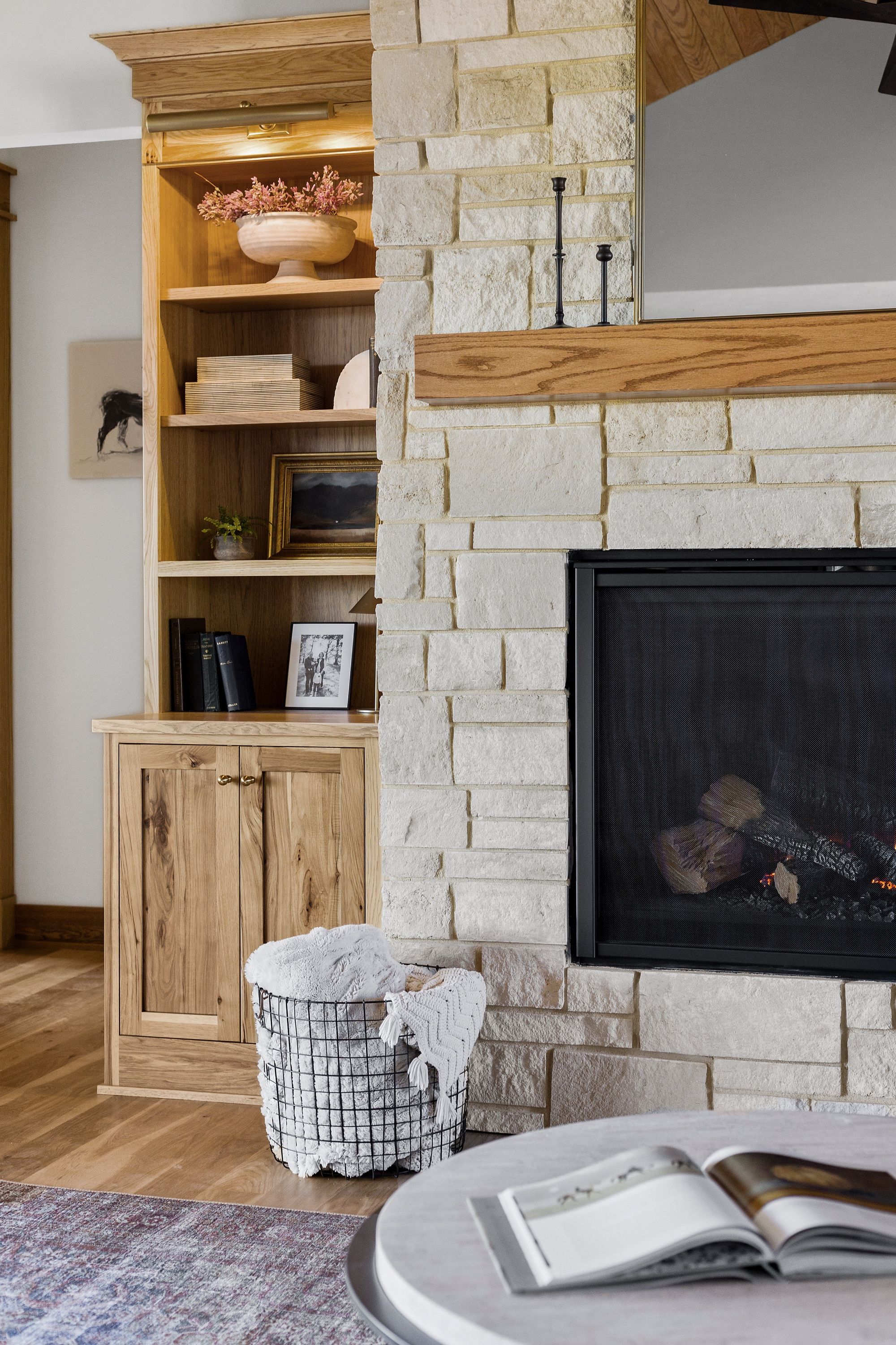 The living room features a stone fireplace with a black firebox, a wooden mantel, and built-in wooden bookshelves. A wire basket with fluffy blankets sits near the fireplace, while an open book rests on the coffee table.