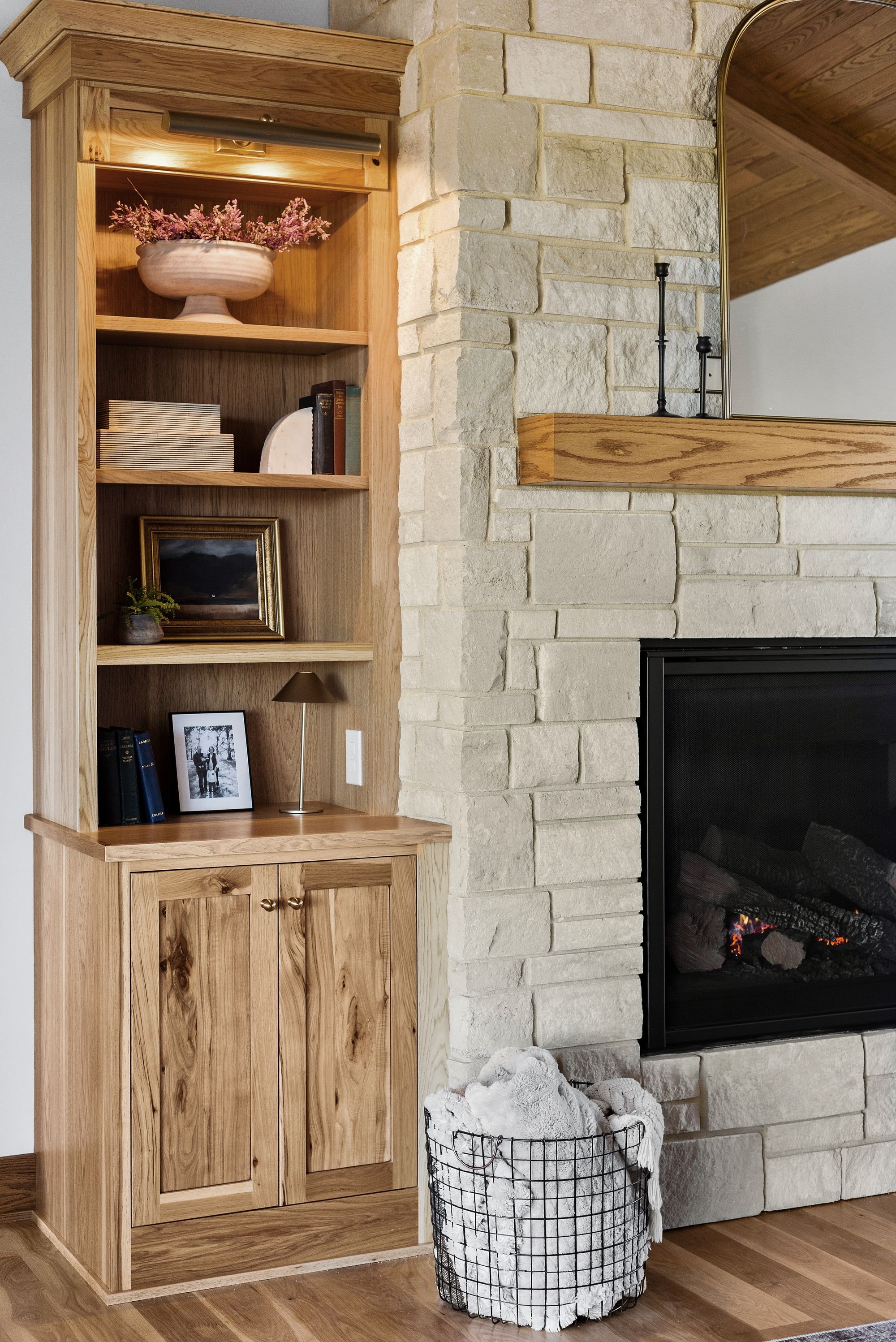 The photo showcases a living room area with a built-in wooden bookshelf next to a stone fireplace with a dark interior and wood mantel. A metal basket with light colored plush blankets sits on a wood floor in front of the fireplace.
