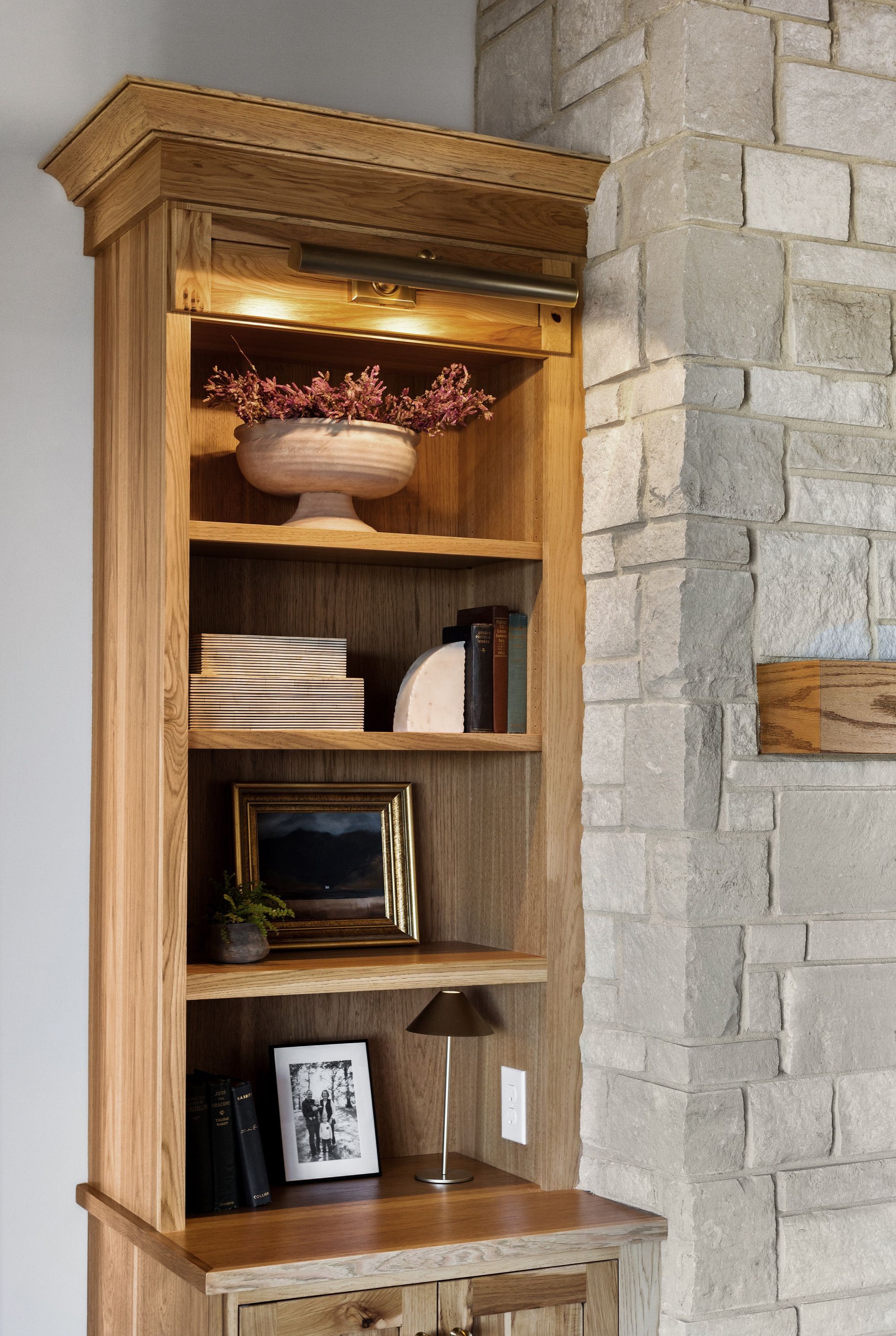 This built-in bookshelf is made of wood and features a light fixture above the top shelf. The shelves display decorative items and books, and the unit is adjacent to a stone wall.