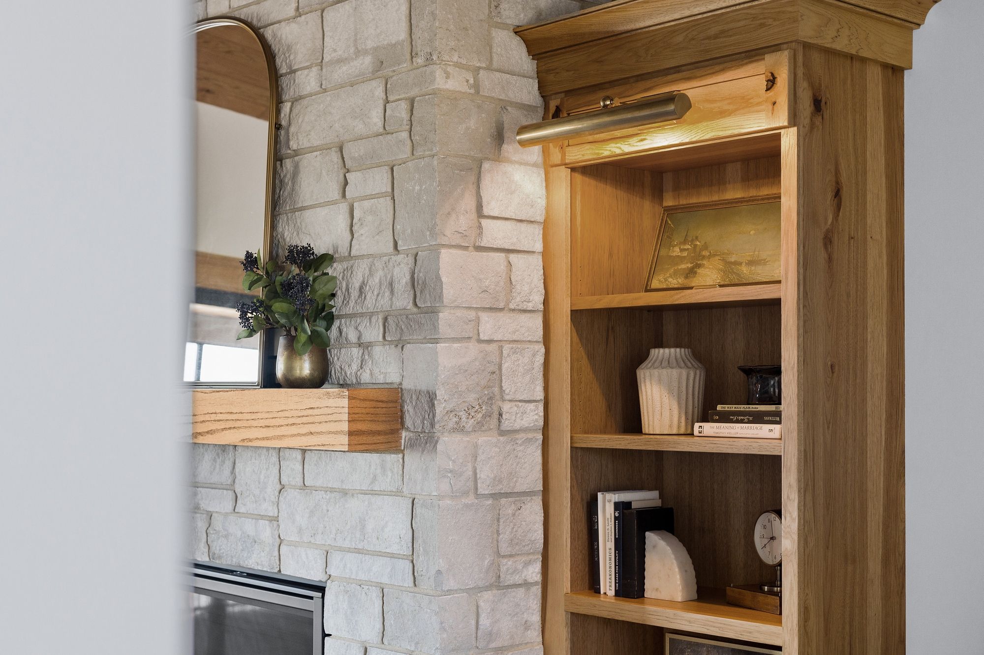 A living room shows a light stone fireplace with a wood mantel and a gold mirror above. Adjacent to the fireplace is a built-in bookshelf made of warm wood.