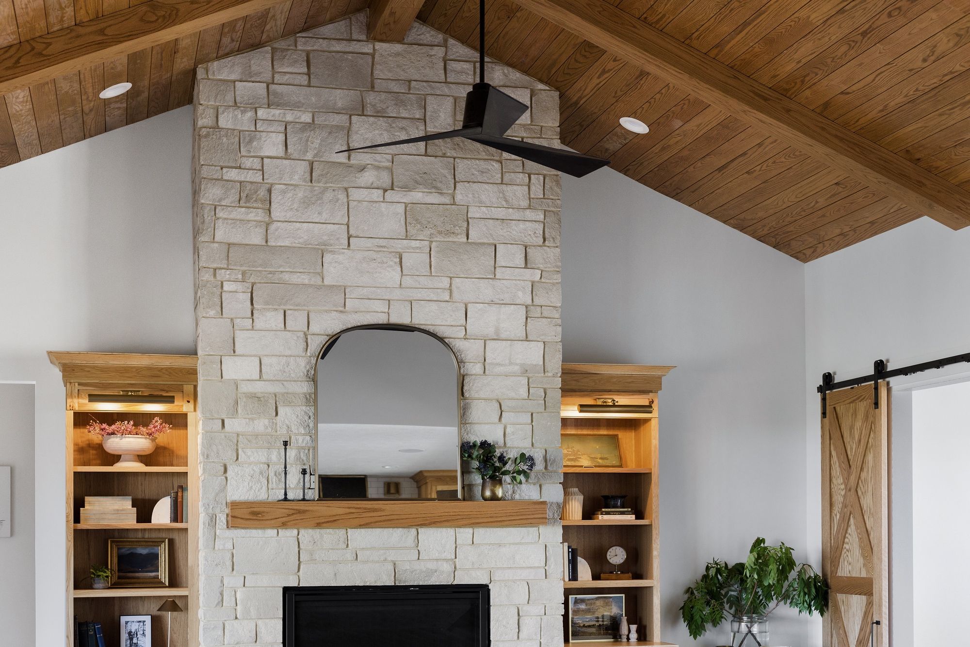 The living room features a stone fireplace with a wooden mantle, built-in wooden shelves, a black ceiling fan, and a light wood planked ceiling with exposed beams. A sliding barn door is located on the right side of the photo.