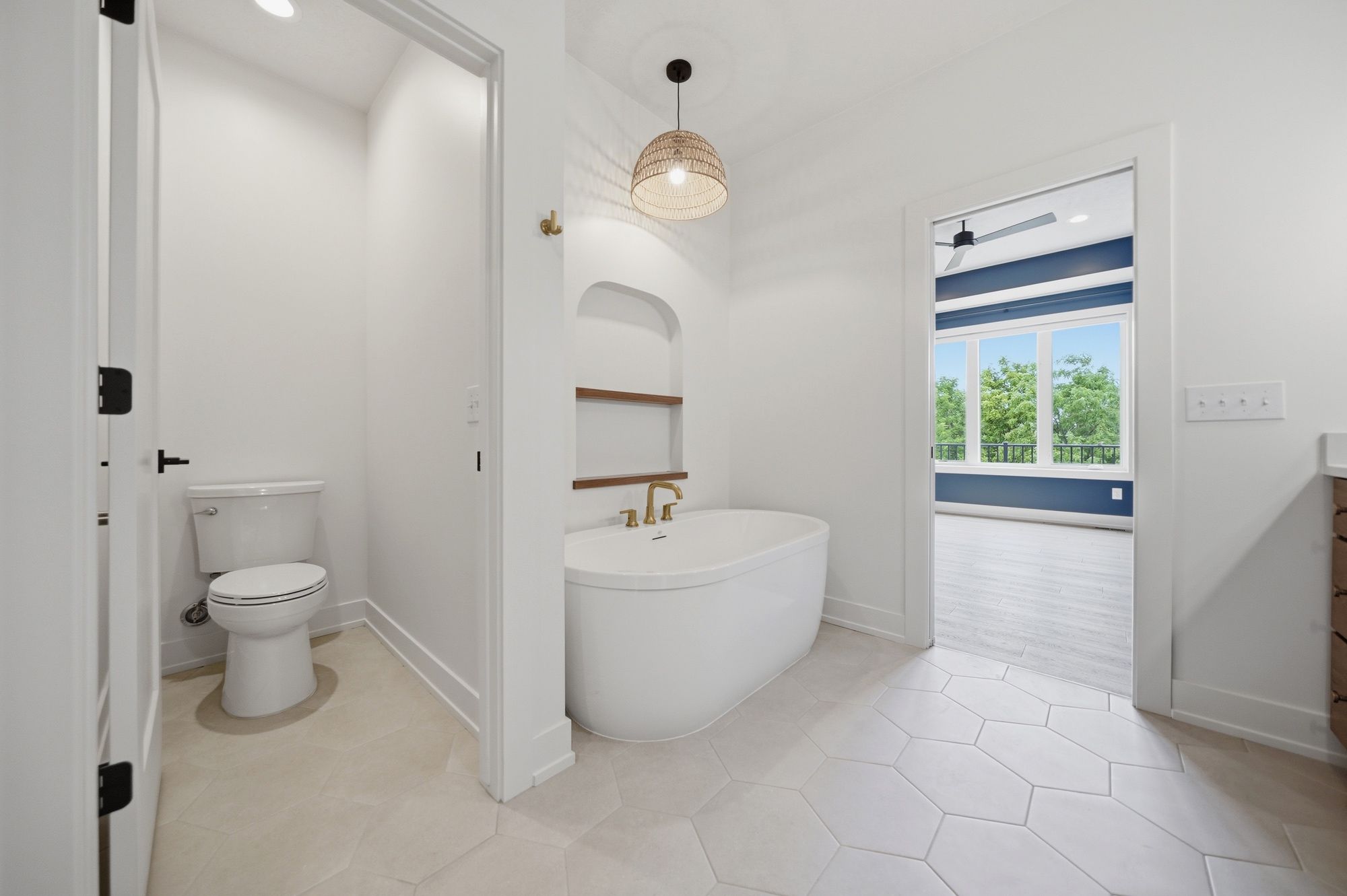 Modern bathroom featuring a freestanding white tub, separate toilet closet, gold fixtures, built-in arched wall niche with wood shelves, and light-colored hexagon floor tile.