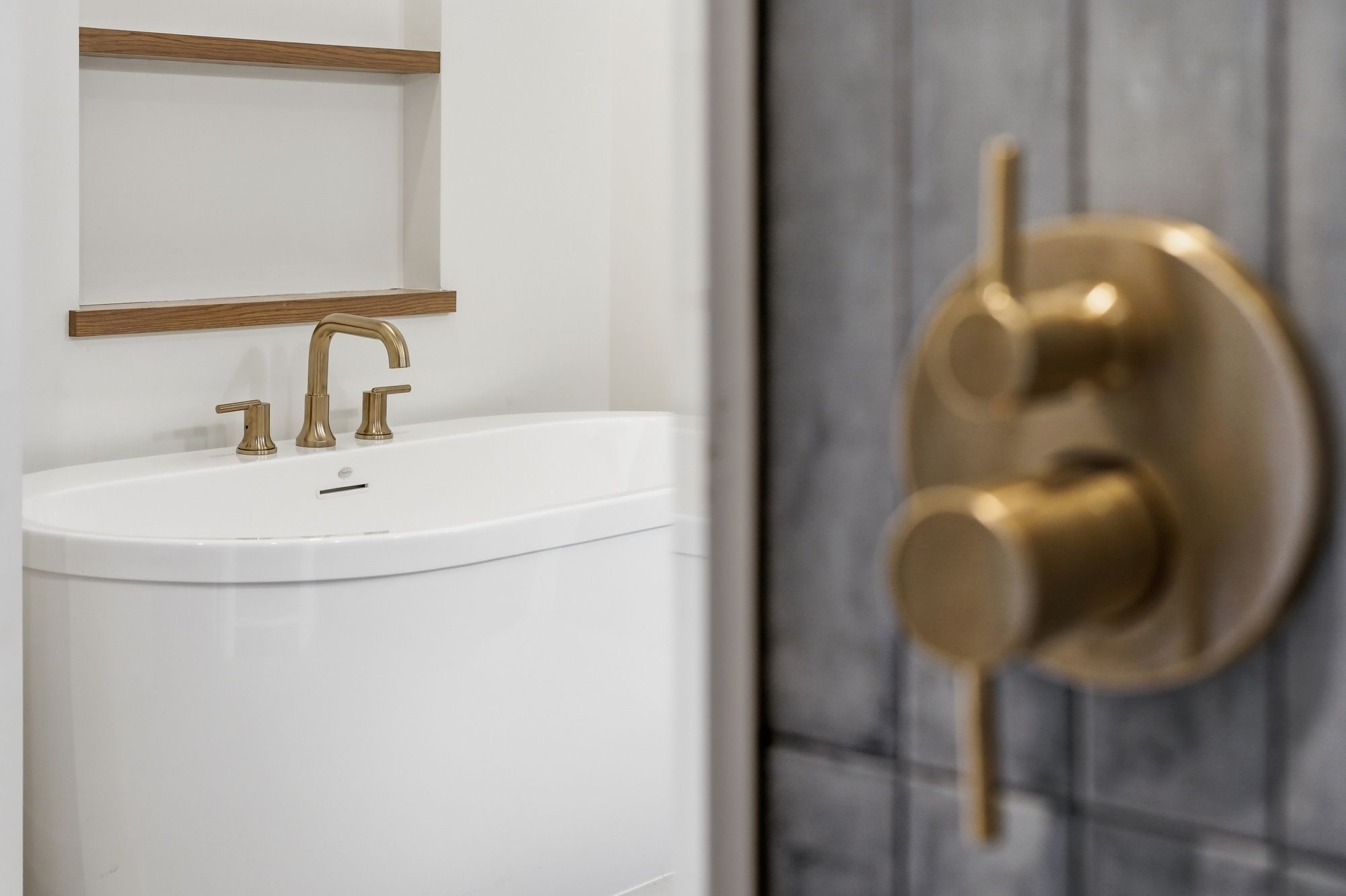 Modern bathroom showcasing a white freestanding soaking tub with a brushed gold faucet, recessed shelving with wood accents, and a close-up of a brushed gold shower control valve on gray tile.
