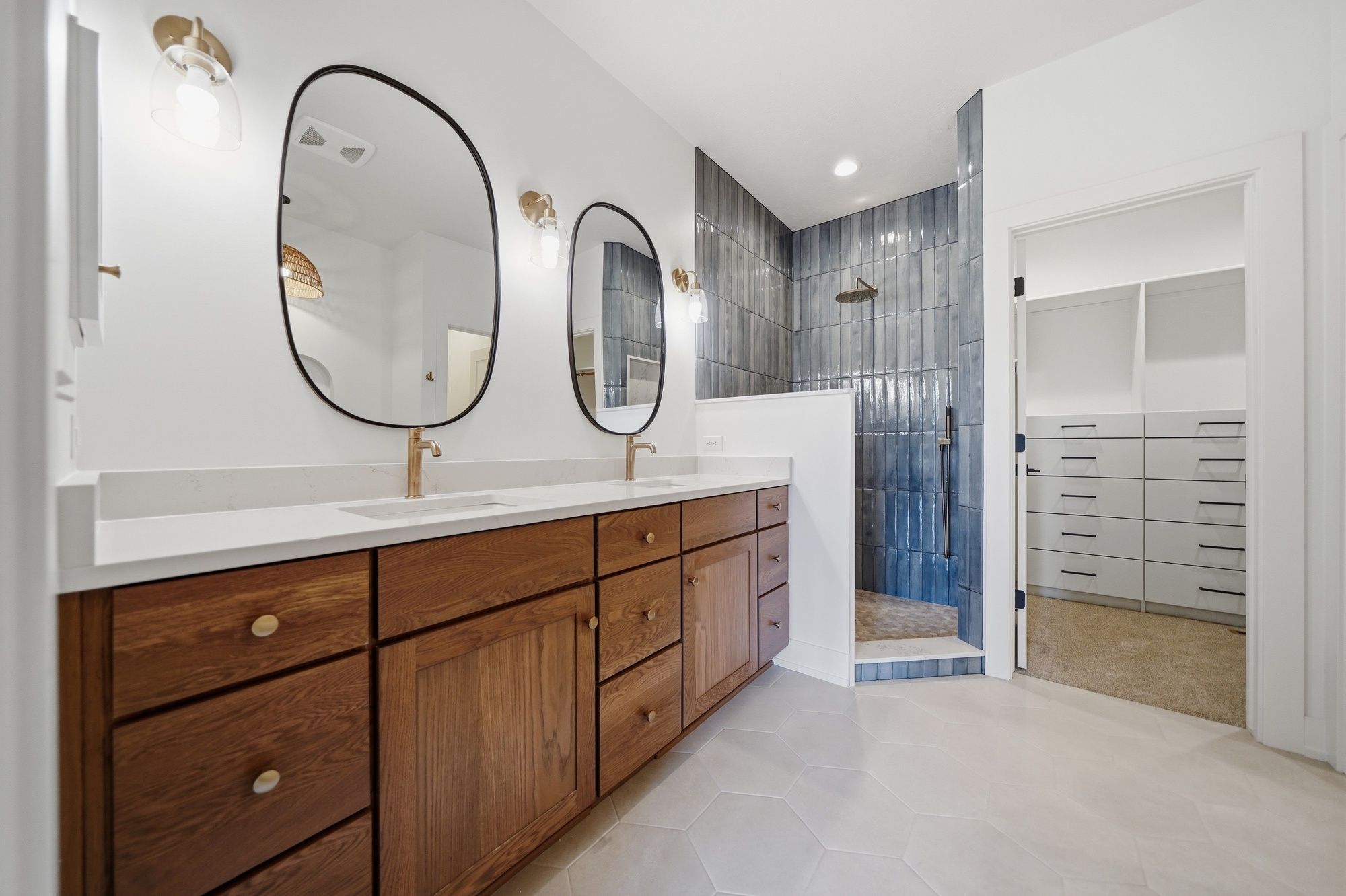 Modern bathroom featuring a warm wood double vanity with gold fixtures, asymmetrical black-framed mirrors, and a walk-in shower with vertical blue tile.