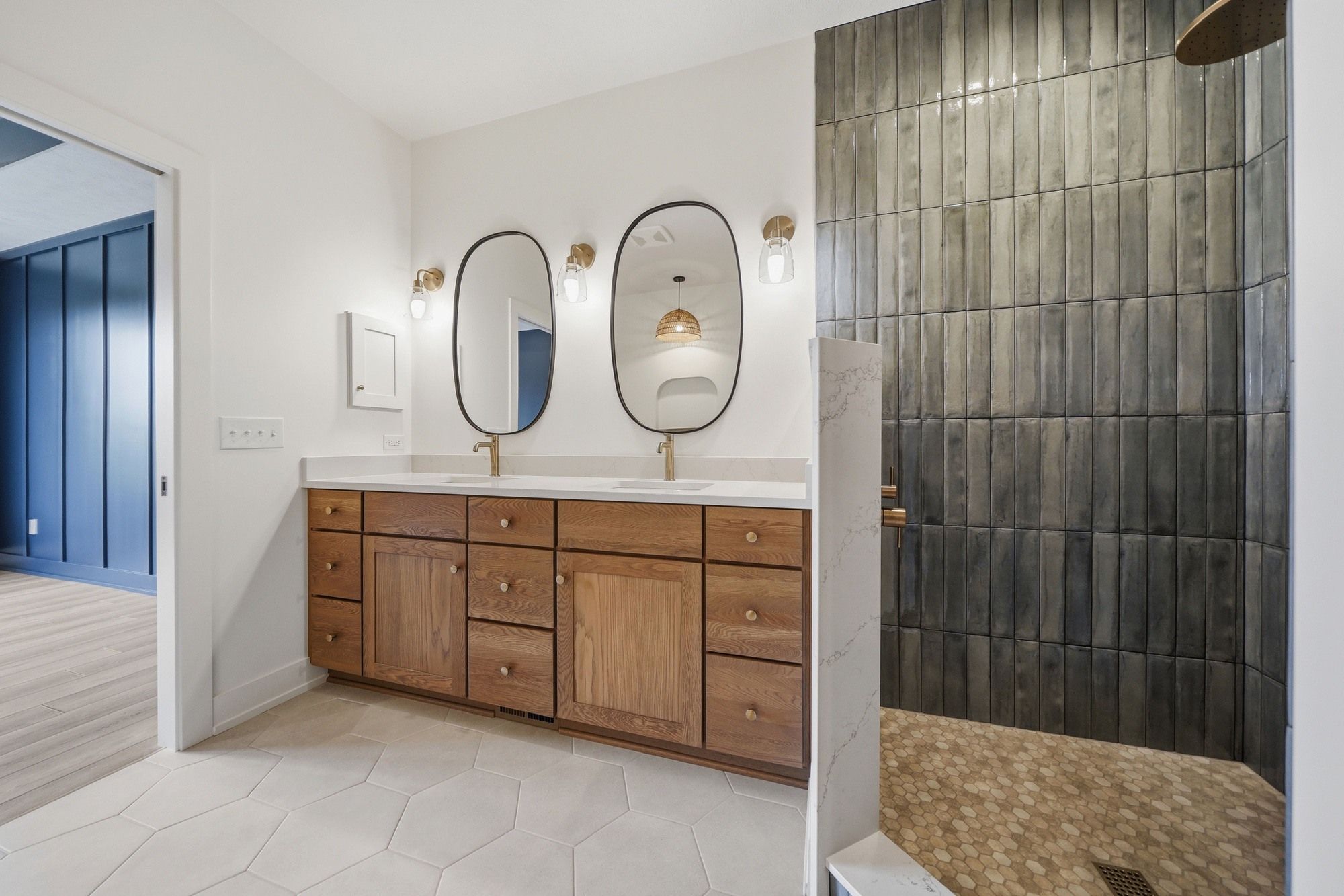 Modern bathroom featuring a wood double vanity with gold fixtures, oval black-framed mirrors, and a walk-in shower with vertical dark gray tiles.