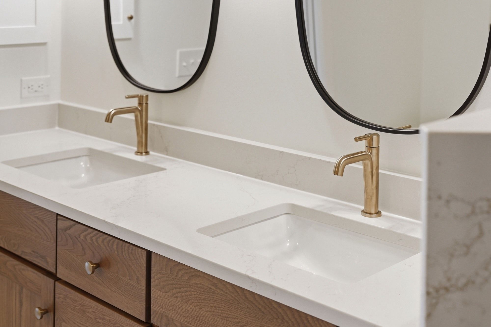Modern bathroom vanity with double undermount sinks, brushed gold faucets, white quartz countertops with light gray veining, wood cabinets, and round black-framed mirrors.