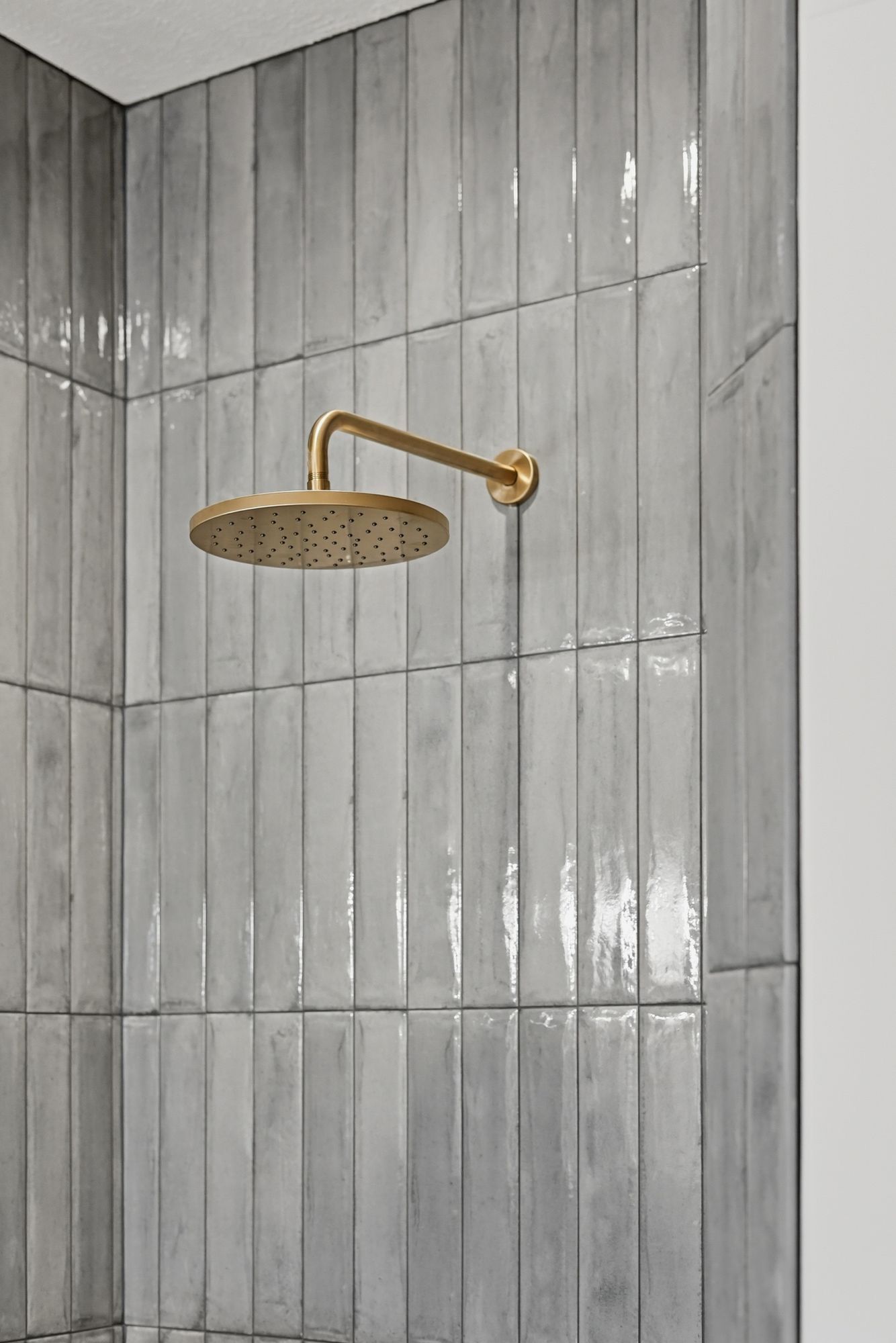 Modern shower featuring glossy, vertically stacked light gray tiles and a brushed gold rain shower head.