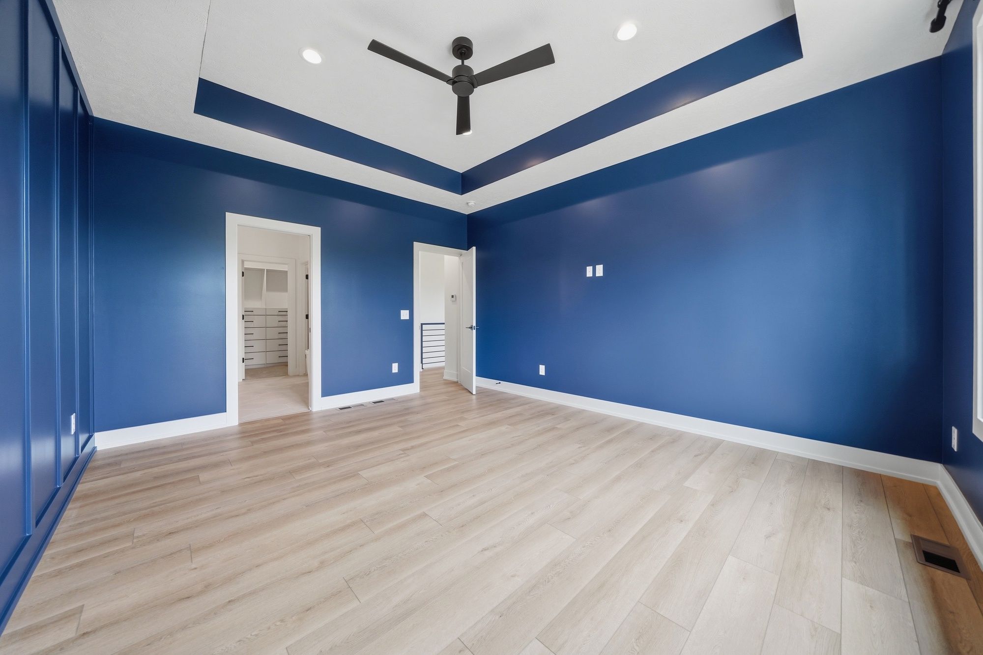 Empty bedroom featuring deep blue walls, a board and batten accent wall, light wood flooring, and a tray ceiling with a matte black ceiling fan.