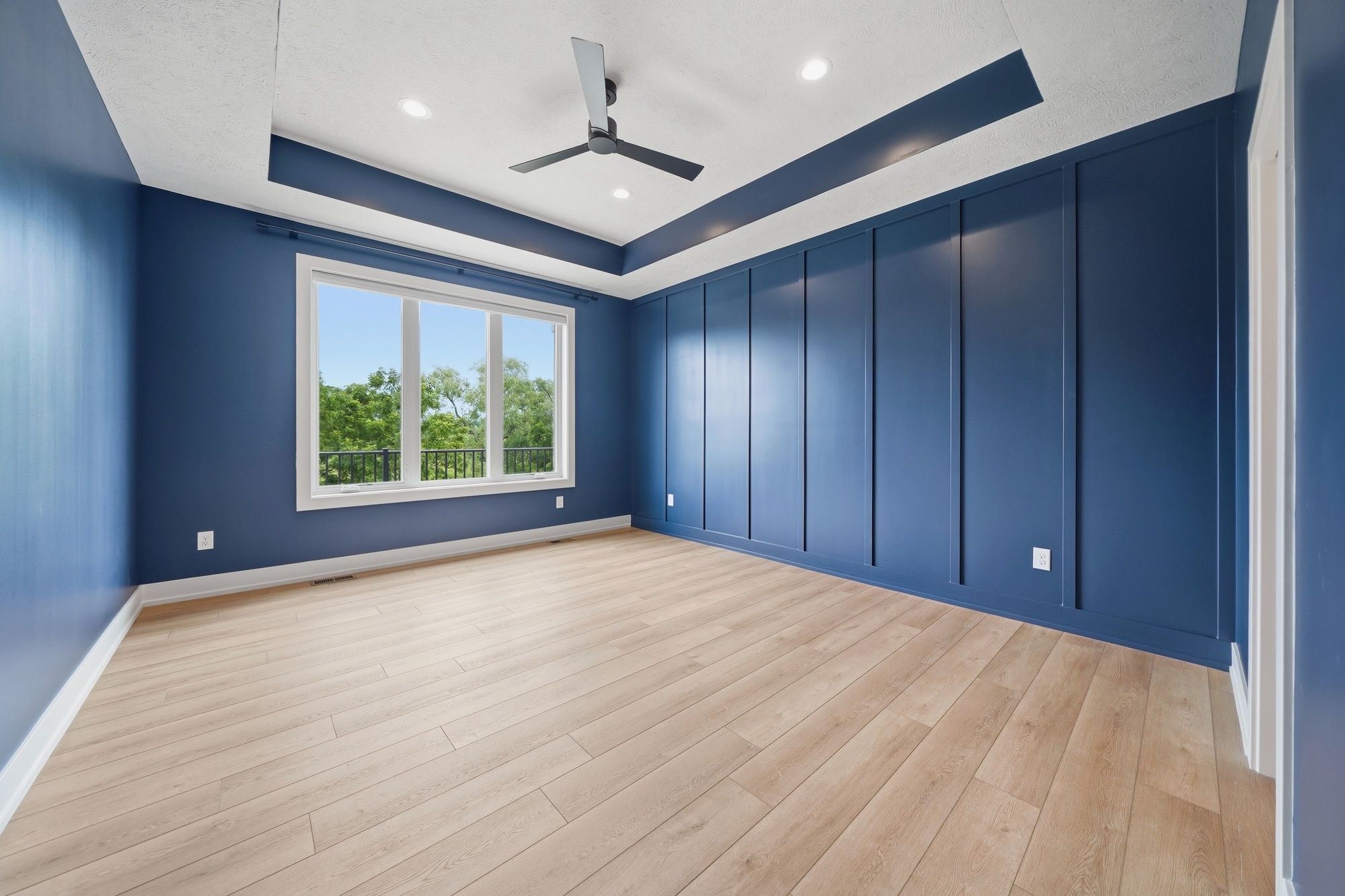 Empty bedroom featuring deep blue walls with a board and batten accent wall, light wood flooring, and a tray ceiling with a black ceiling fan.