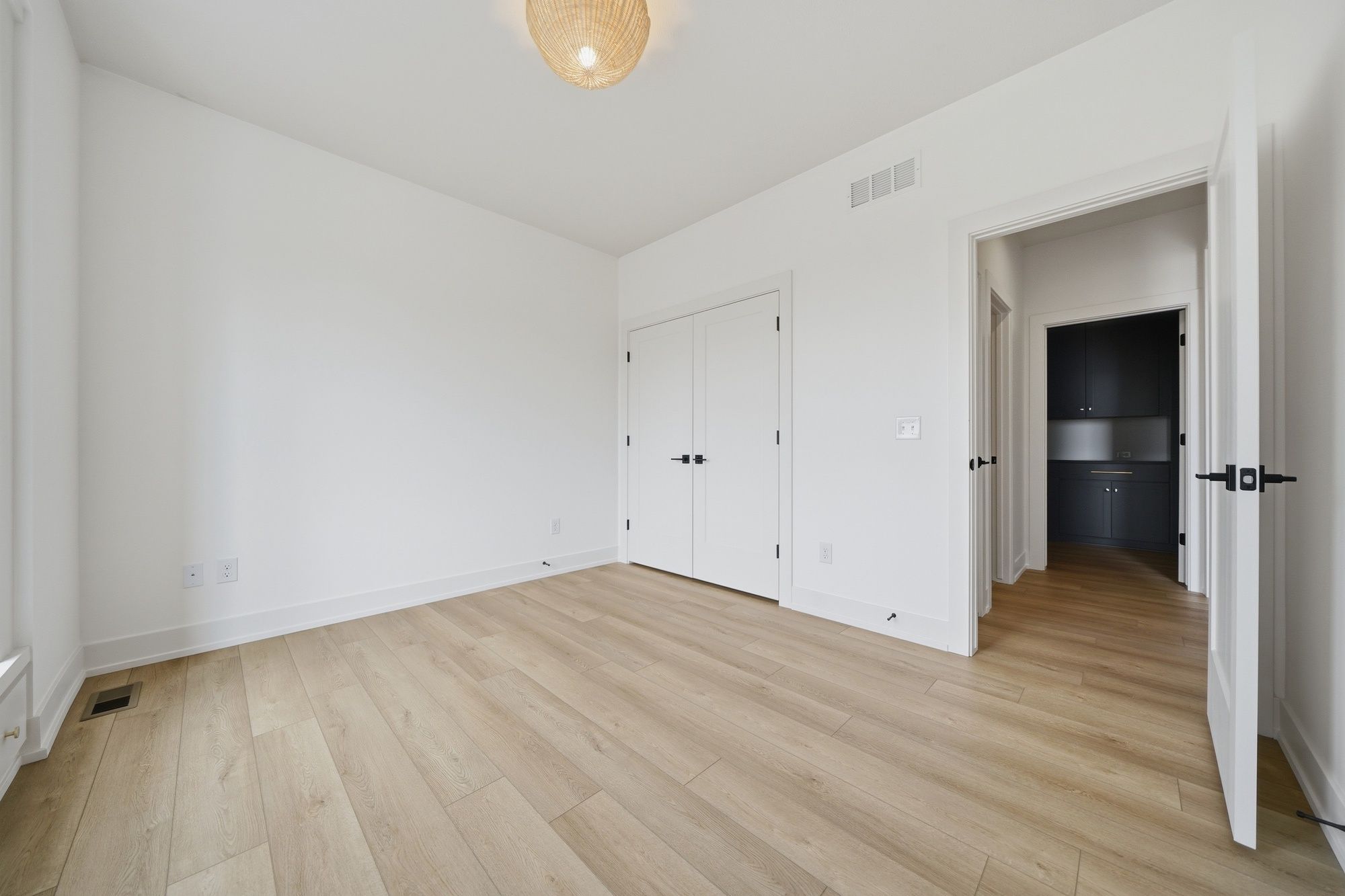 Empty bedroom featuring light wood-look flooring, crisp white walls, a double-door closet with black hardware, and a woven dome ceiling light.