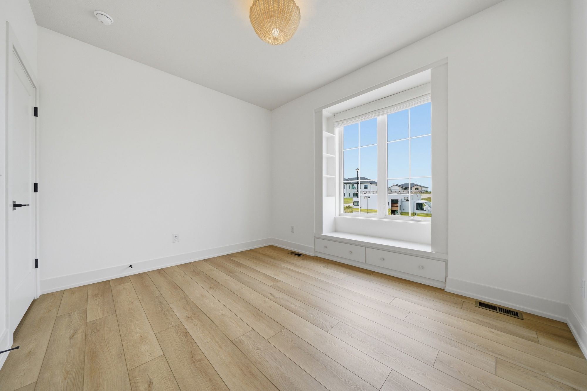 Empty bedroom featuring light wood floors, clean white walls, and a large window with a built-in window seat, storage drawers, and shelving.