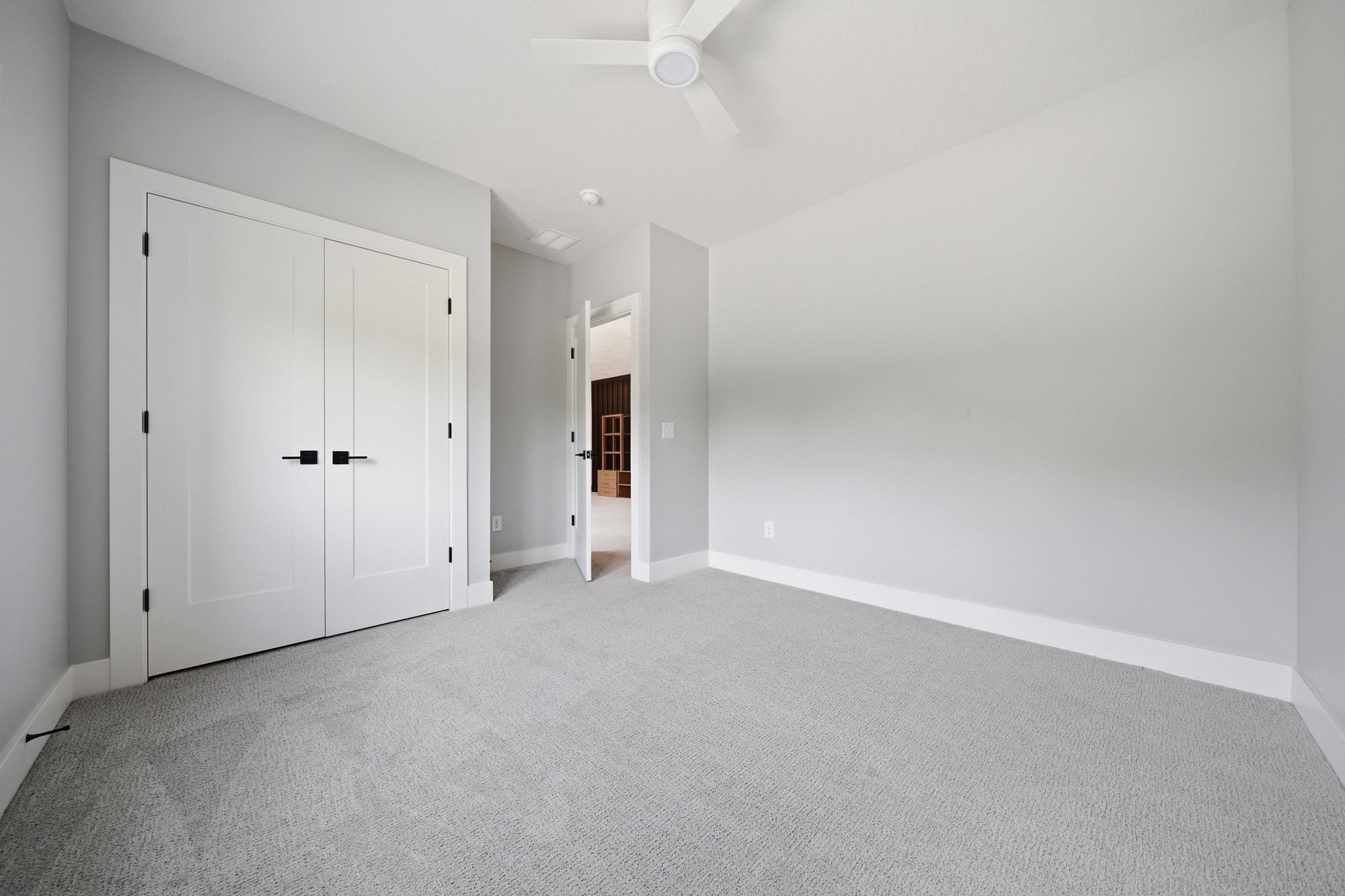 Empty modern bedroom with light gray carpet, soft gray walls, double closet doors with black handles, and a white ceiling fan.