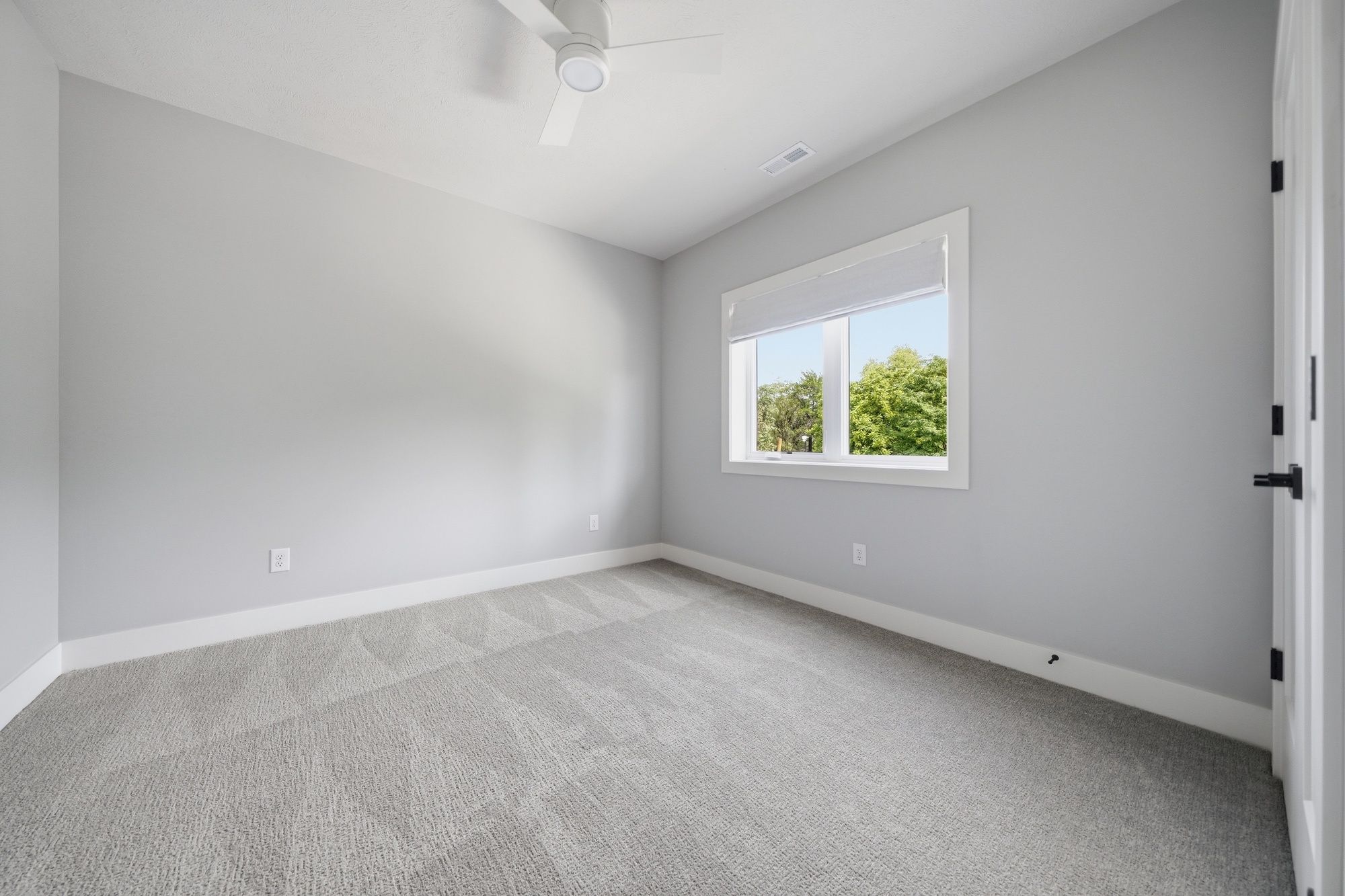 Empty modern bedroom featuring light gray walls, plush gray carpeting, a white ceiling fan, and a single window showing outdoor trees.
