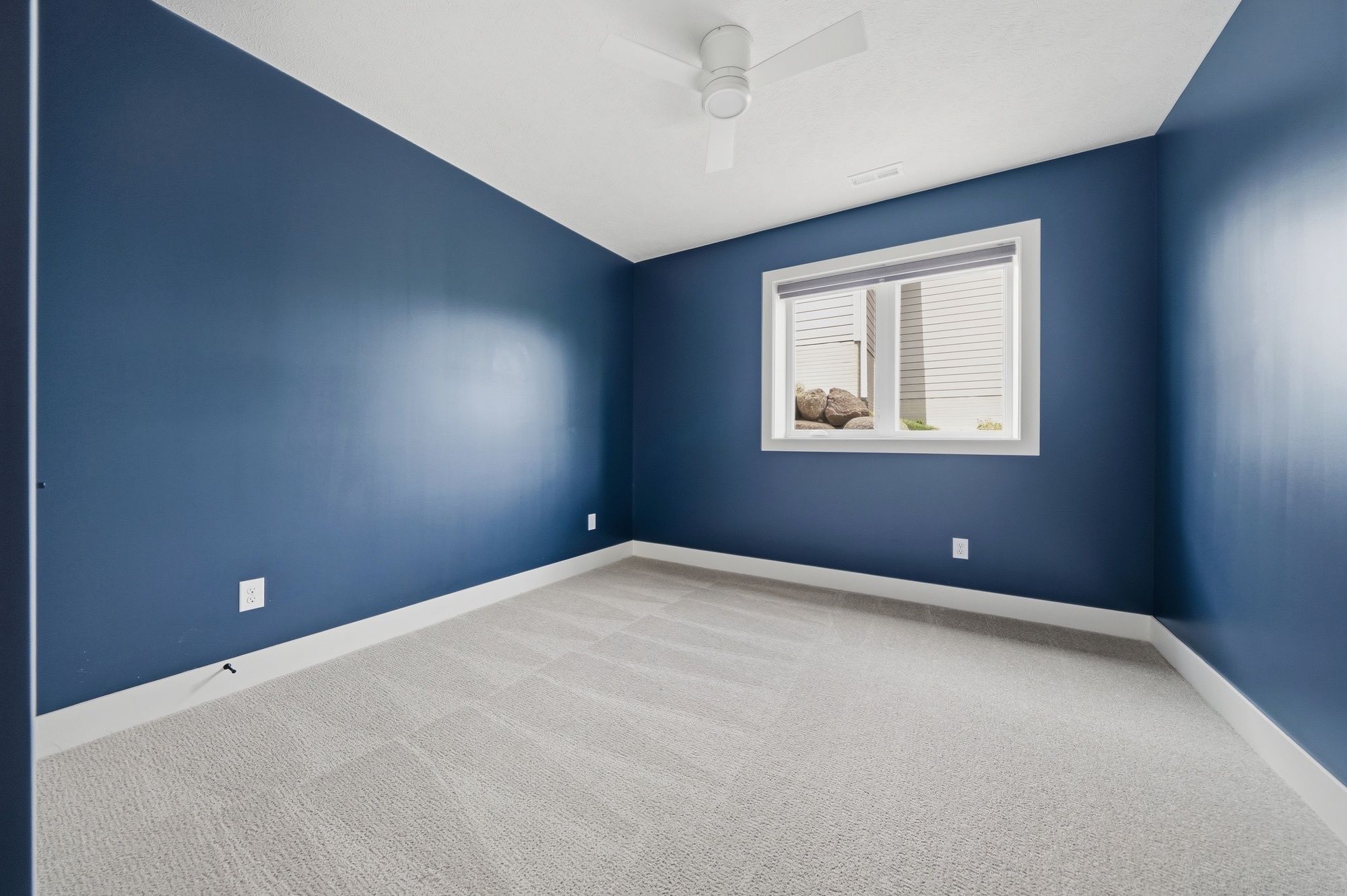 Empty bedroom featuring deep blue walls, light gray carpeting, a white ceiling fan, and white baseboards.