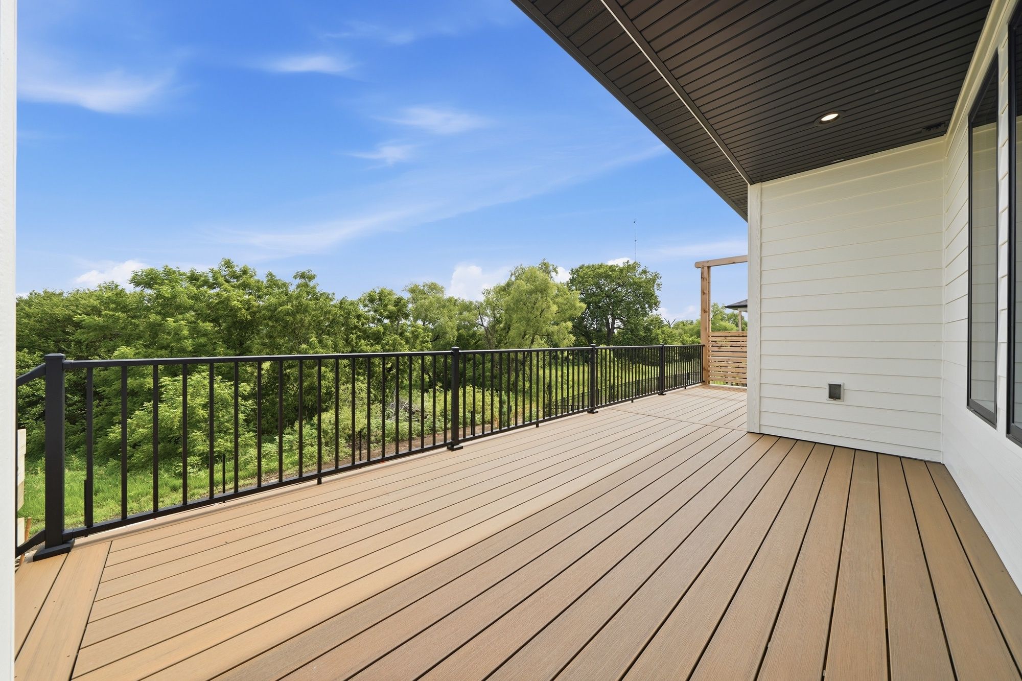 Spacious covered deck featuring warm brown composite decking, sleek black metal railings, and a dark paneled soffit ceiling overlooking a lush green treeline.