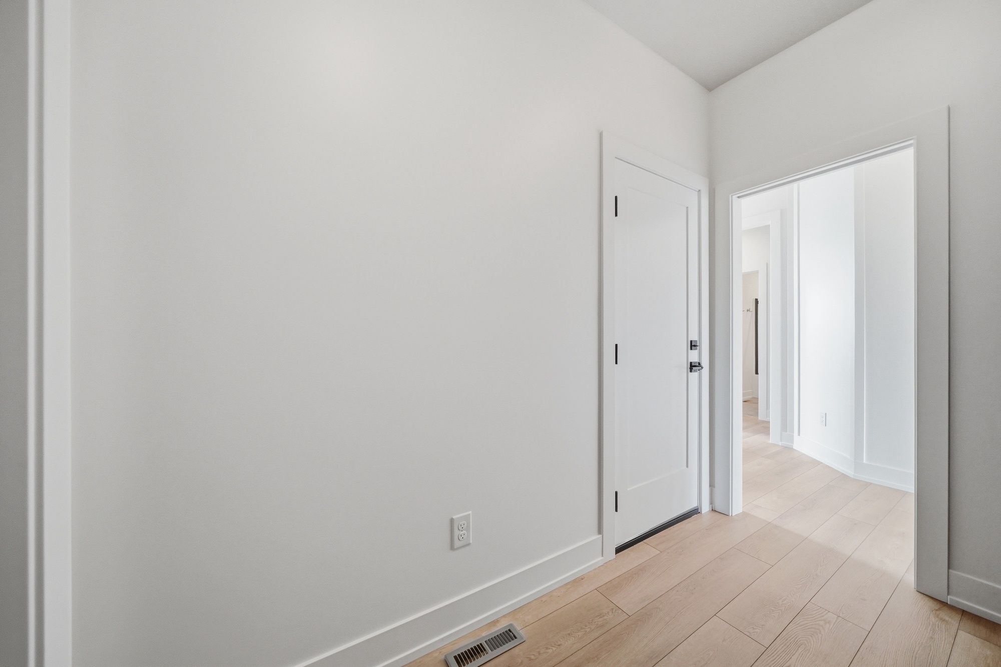 Bright hallway with light wood floors, crisp white walls, and a white door featuring black hardware.