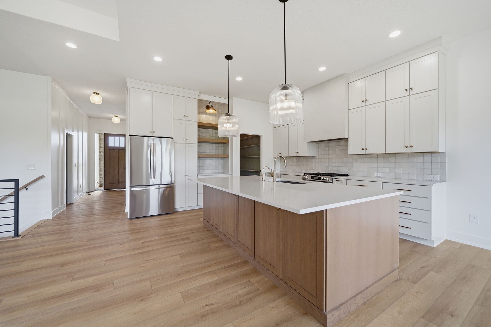 Modern open-concept kitchen featuring white cabinets, a large wood-toned island with white quartz countertops, stainless steel appliances, and ribbed glass pendant lights.