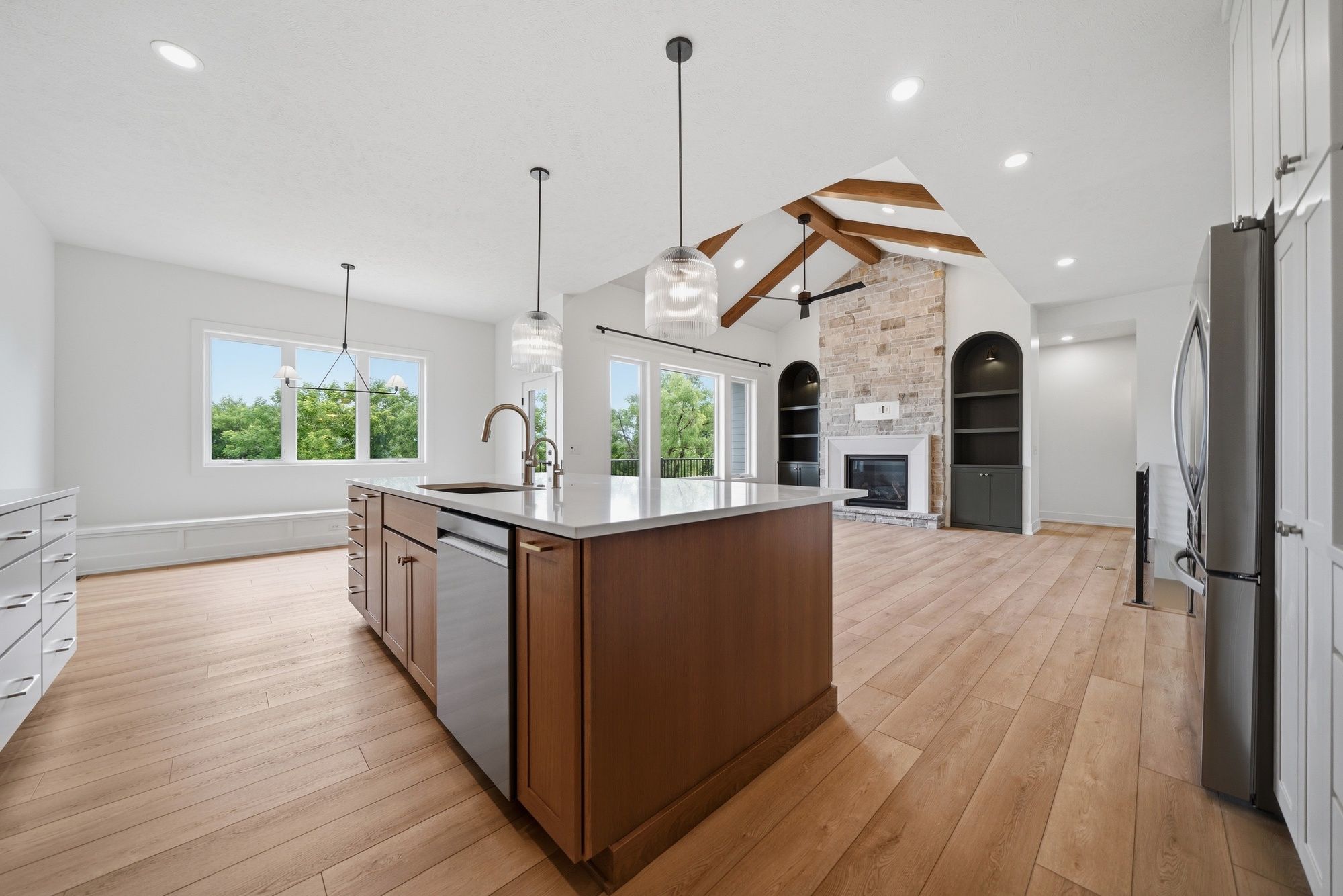 Open-concept kitchen featuring a warm wood island with white quartz countertops and a stainless steel dishwasher, looking into a living area with a stone fireplace and vaulted ceiling with exposed wood beams.