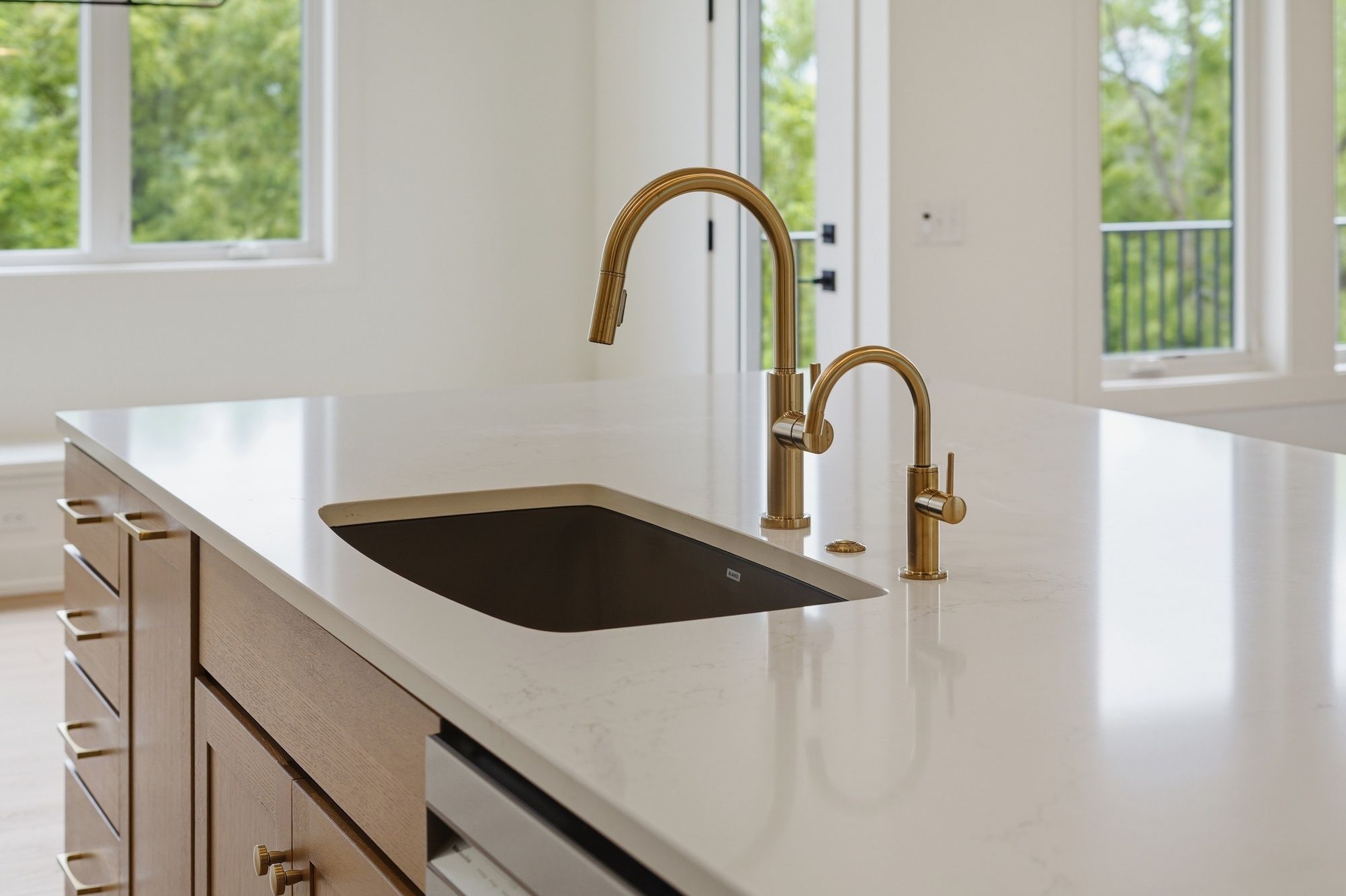 Modern kitchen island featuring a white quartz countertop with a dark undermount sink, brushed gold gooseneck faucets, and light wood cabinetry.