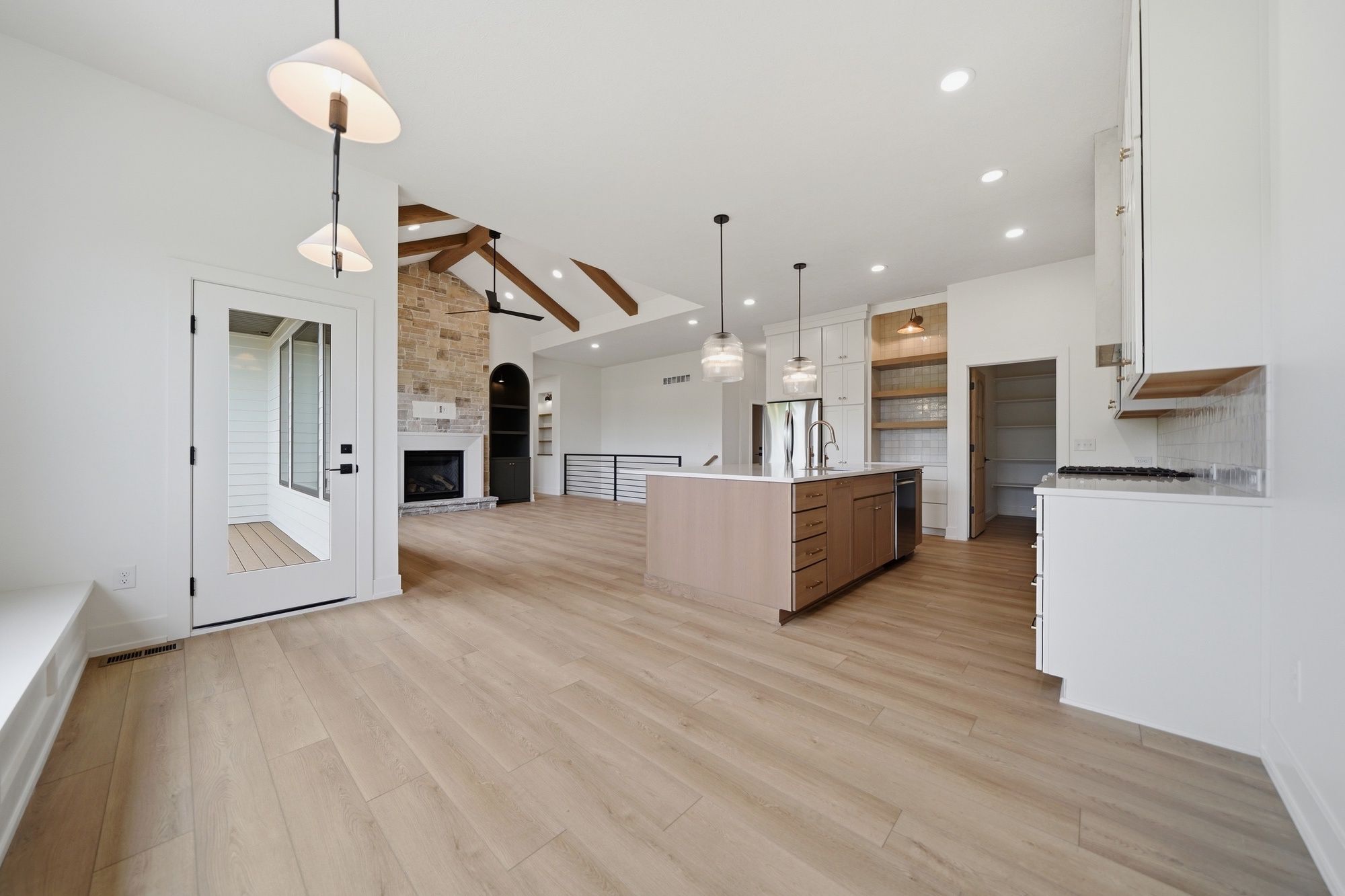 Open-concept kitchen and living area featuring light wood floors, a central wood island with white countertops, and a stone fireplace under a vaulted ceiling with exposed wood beams.