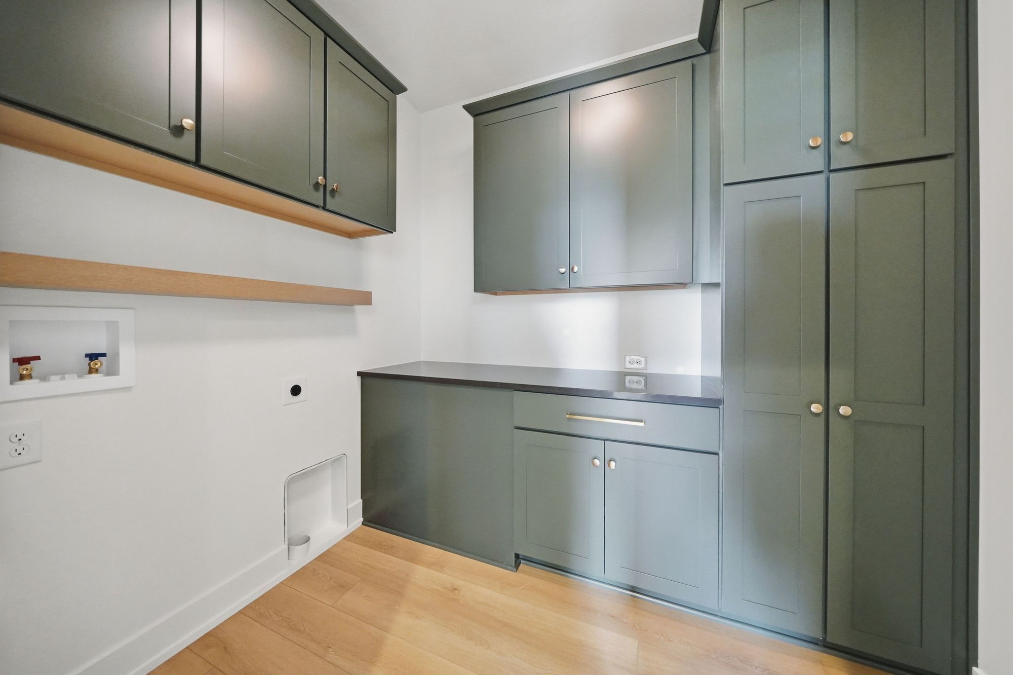Laundry room featuring sage green cabinetry with gold hardware, a dark countertop, light hardwood floors, and empty washer and dryer hookups.