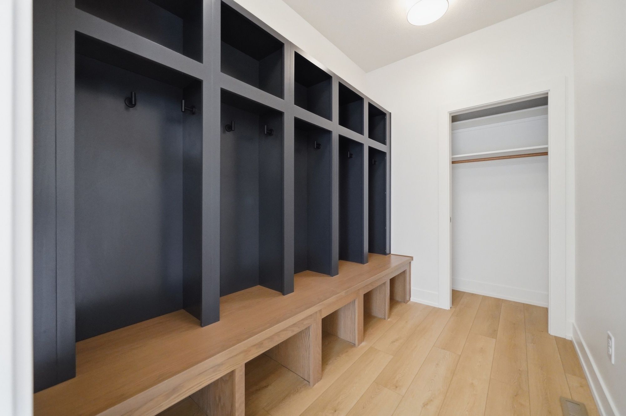 Modern mudroom featuring charcoal black built-in lockers with hooks and a warm wood bench over open cubbies, alongside light wood floors and an open closet.