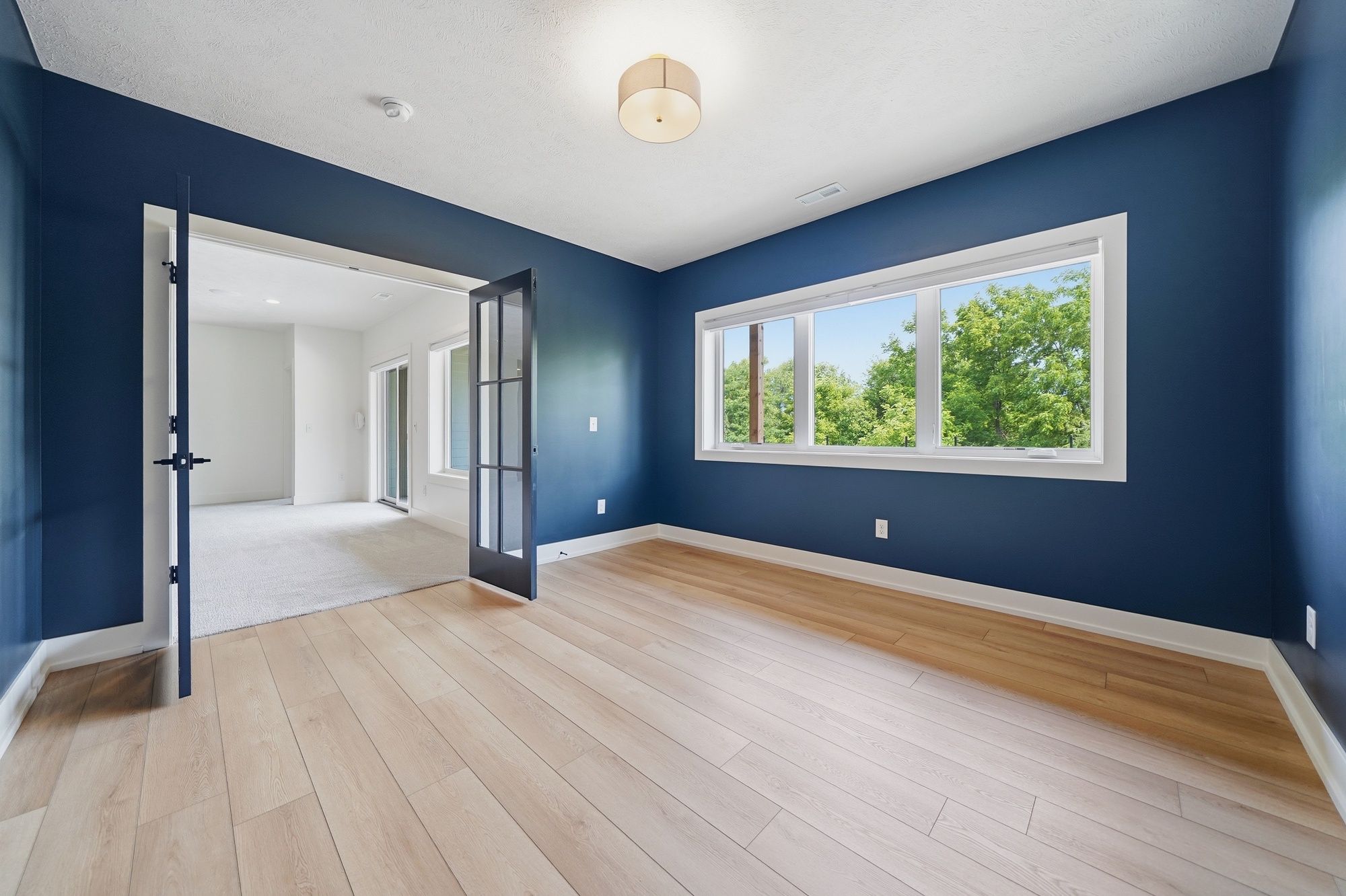 Home office featuring navy blue walls, light hardwood floors, and a large window looking out to trees, with dark-framed French doors opening to an adjacent carpeted area.