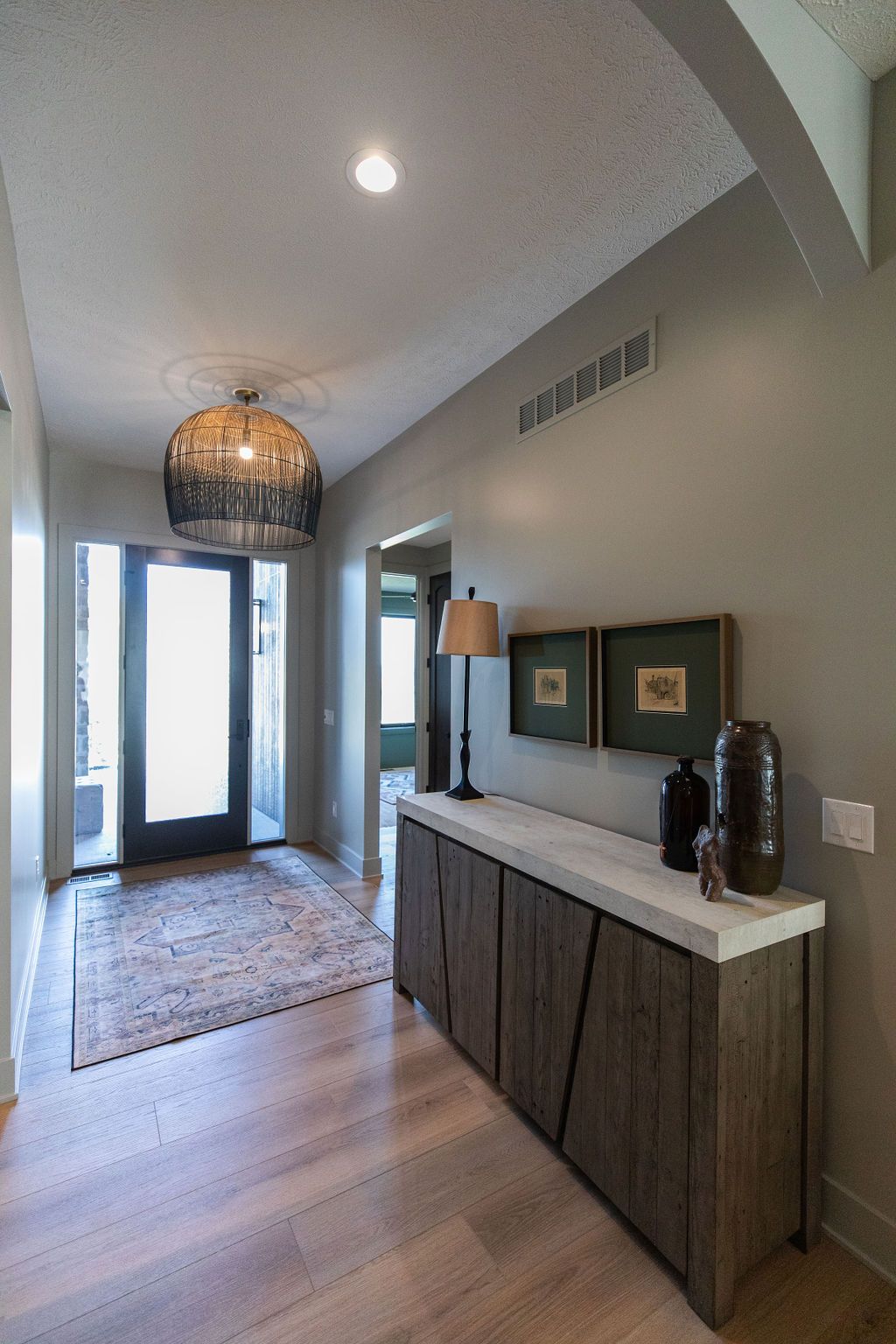 Entryway with light wood floors, a patterned rug, and a black-framed glass front door. A rustic sideboard with a concrete top holds a lamp, framed artwork, and decorative vases.