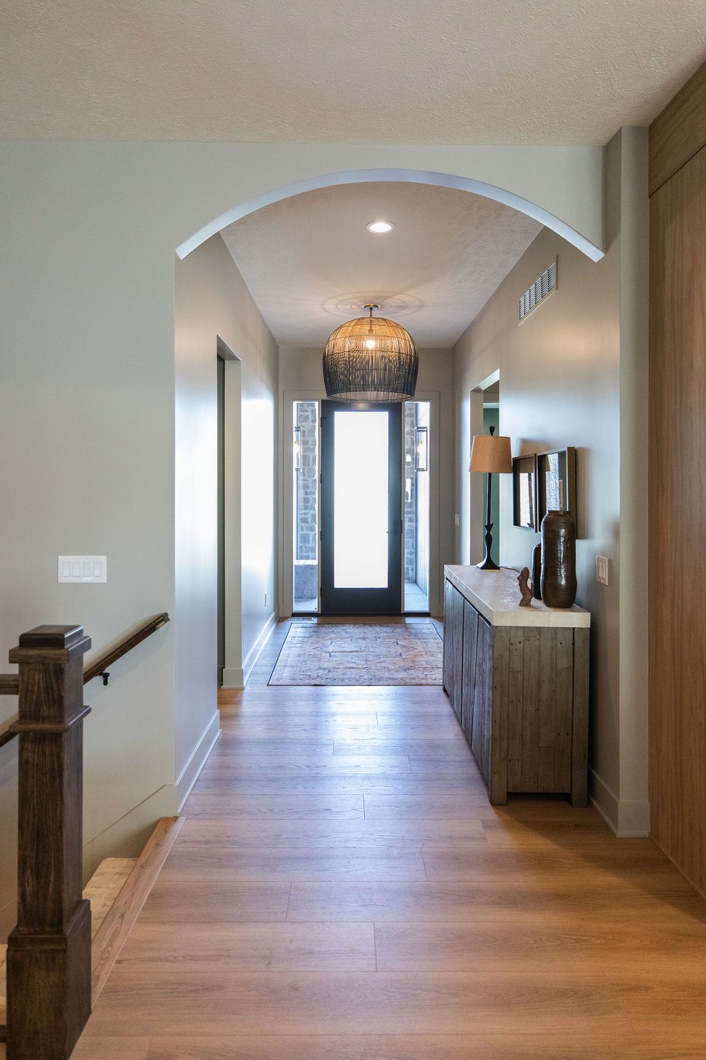 Entryway with light wood-look floors and a large dark wicker pendant light. The space features an arched ceiling detail and leads to a glass front door with sidelights.