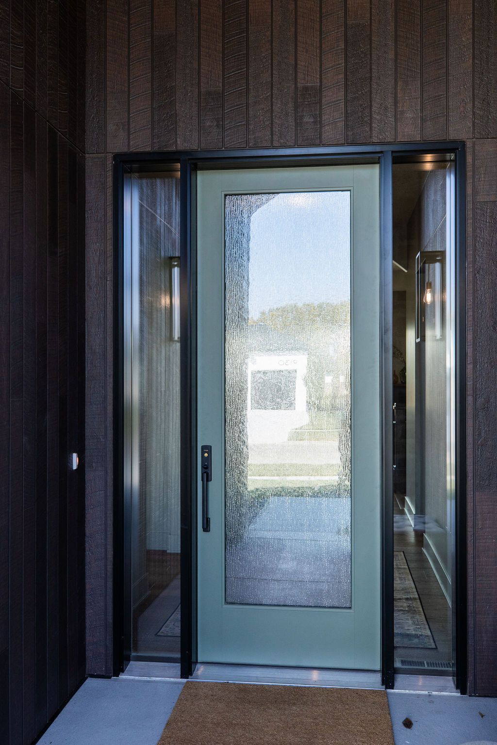 Exterior entryway featuring a modern light green front door with reeded glass and sidelights. The facade is clad in stained wood panels.