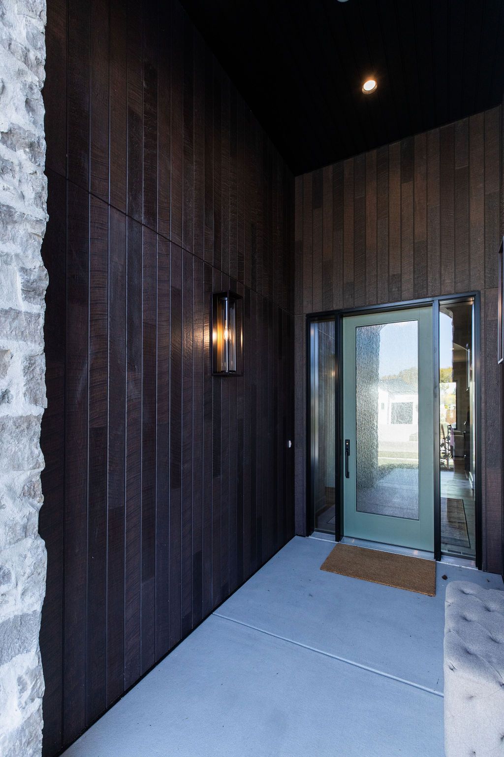 Exterior entryway features a modern door and wall-mounted light fixture. The entryway features dark wood paneling with gray stone accents and a concrete walkway.