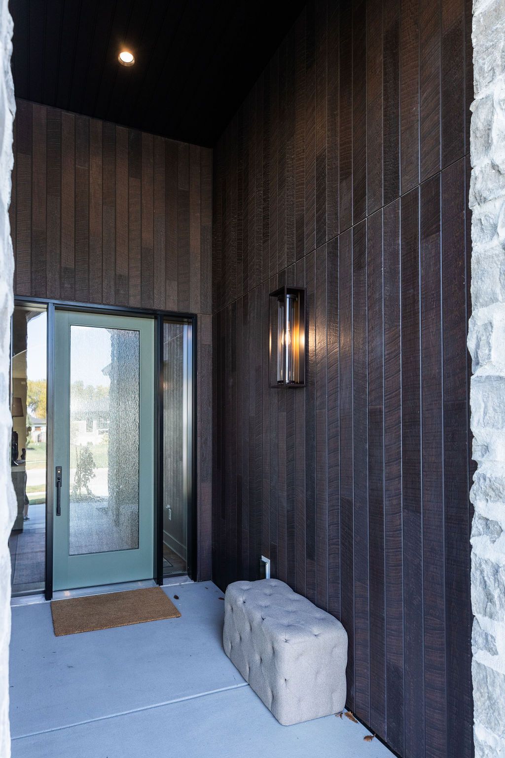 Exterior entryway featuring a wood-paneled wall with a modern light fixture, a light gray cushioned bench, and a green-framed glass door with a brown doormat. Stone pillars flank the entrance, adding texture and contrast to the wooden facade.