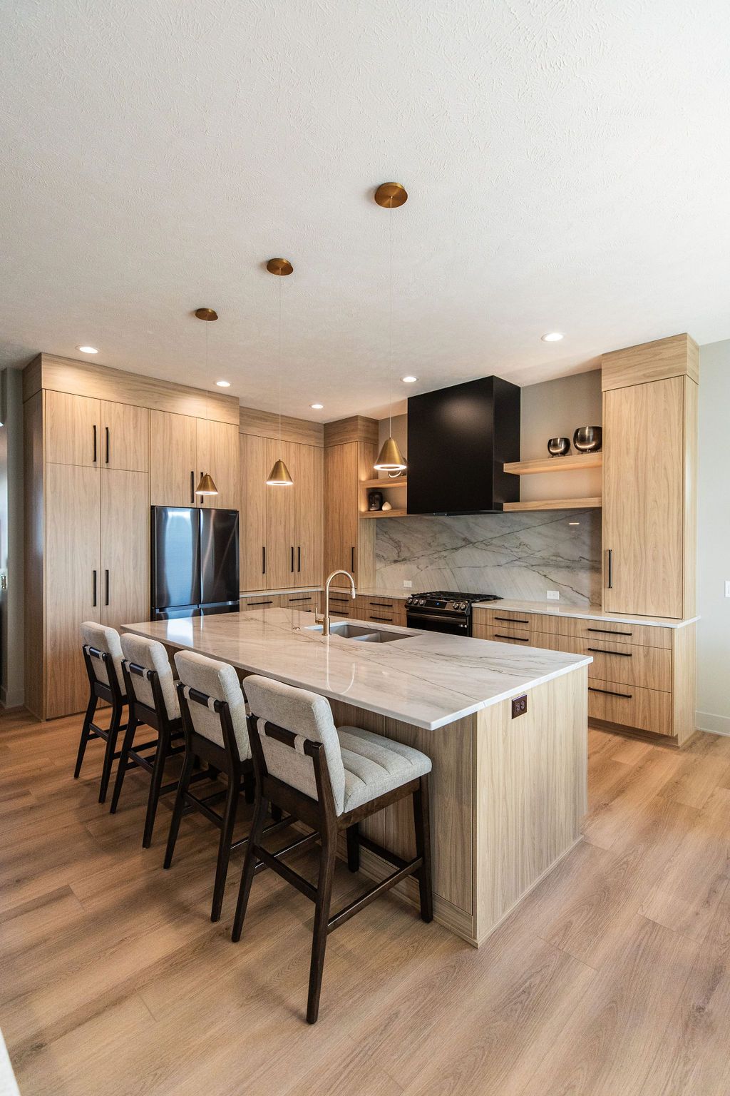 Modern kitchen features light wood cabinetry, a marble-topped island with seating, and stainless steel appliances. Pendant lighting and recessed lighting illuminate the space, which has wood floors and a black range hood.