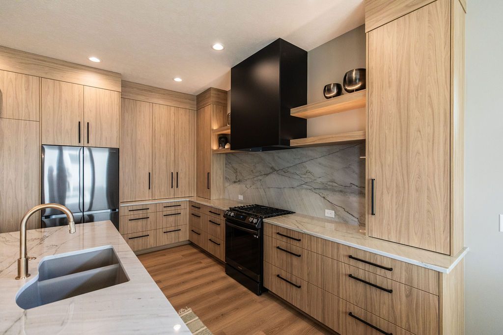 Modern kitchen with light wood cabinetry, marble countertops and backsplash, and stainless steel appliances. Features a black range hood and floating wood shelves.