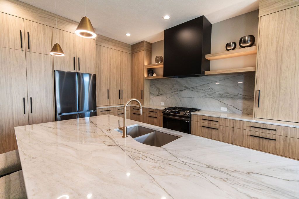 Modern kitchen featuring light wood cabinetry, a black range hood, and a large white marble island with a stainless steel sink and gold faucet. The stainless steel refrigerator complements the cabinets.