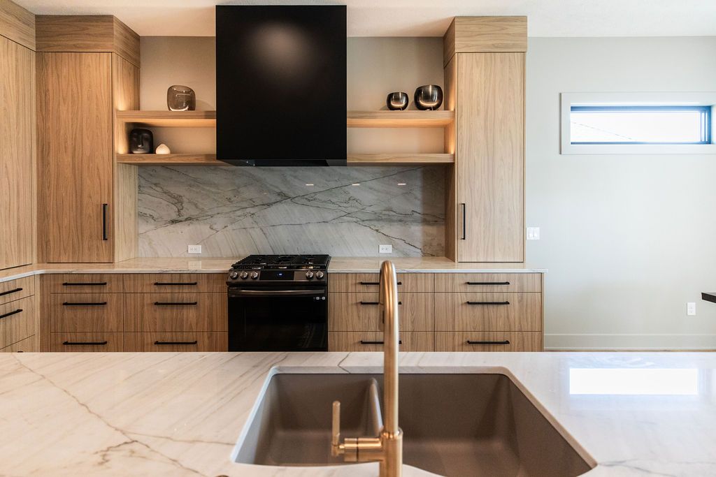 Modern kitchen featuring light wood cabinets with black hardware, marble countertops and backsplash, and a black range hood. The kitchen includes a double sink with a gold faucet, open shelving with decorative items, and stainless steel appliances.