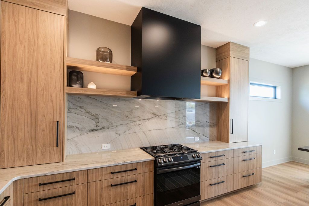 Modern kitchen featuring light wood cabinetry with black hardware, a marble backsplash, and a black range hood. The kitchen also has light-colored countertops and open shelving with decorative items.
